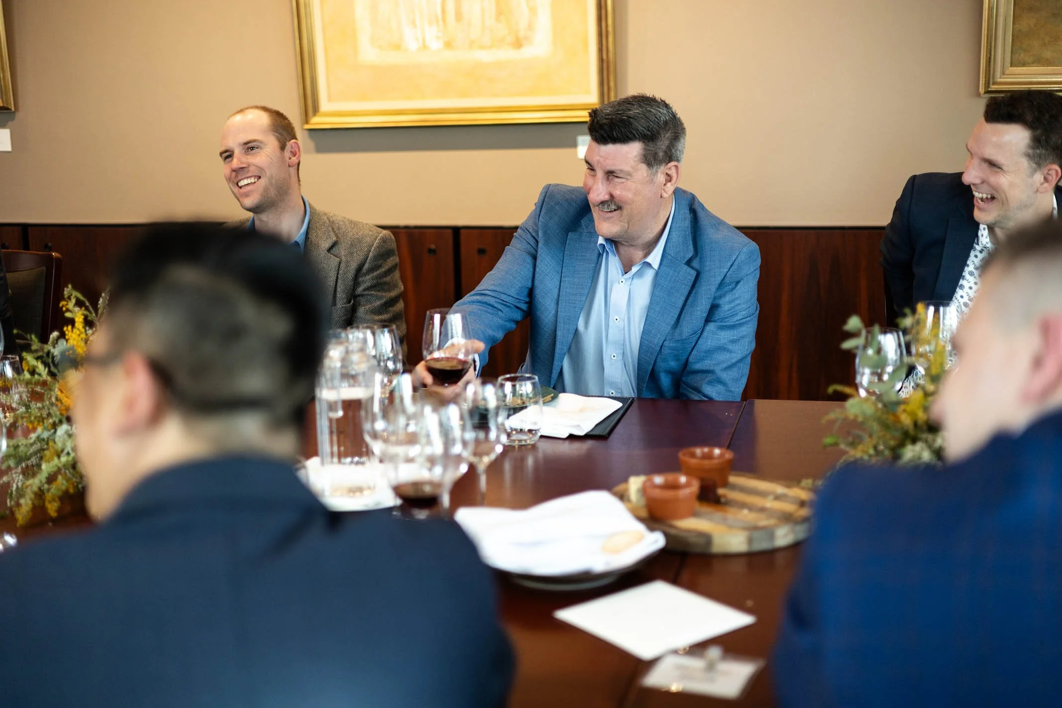 A group of men in business attire sitting around a table, laughing and enjoying a meal during a social gathering or business meeting.