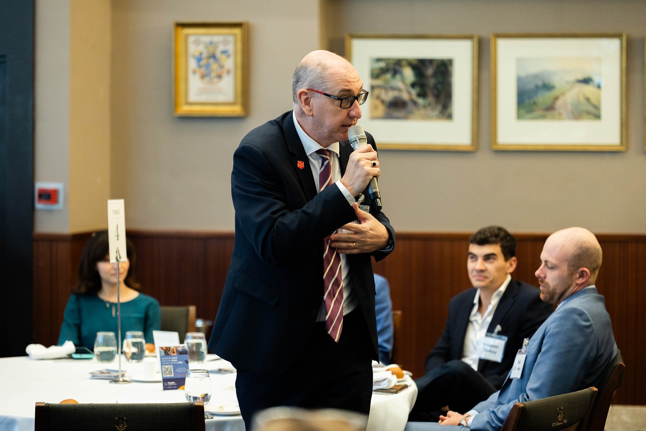 A man in a suit and striped tie holding a microphone and speaking at a formal event or conference, with three seated attendees and framed artwork on the wall in the background.