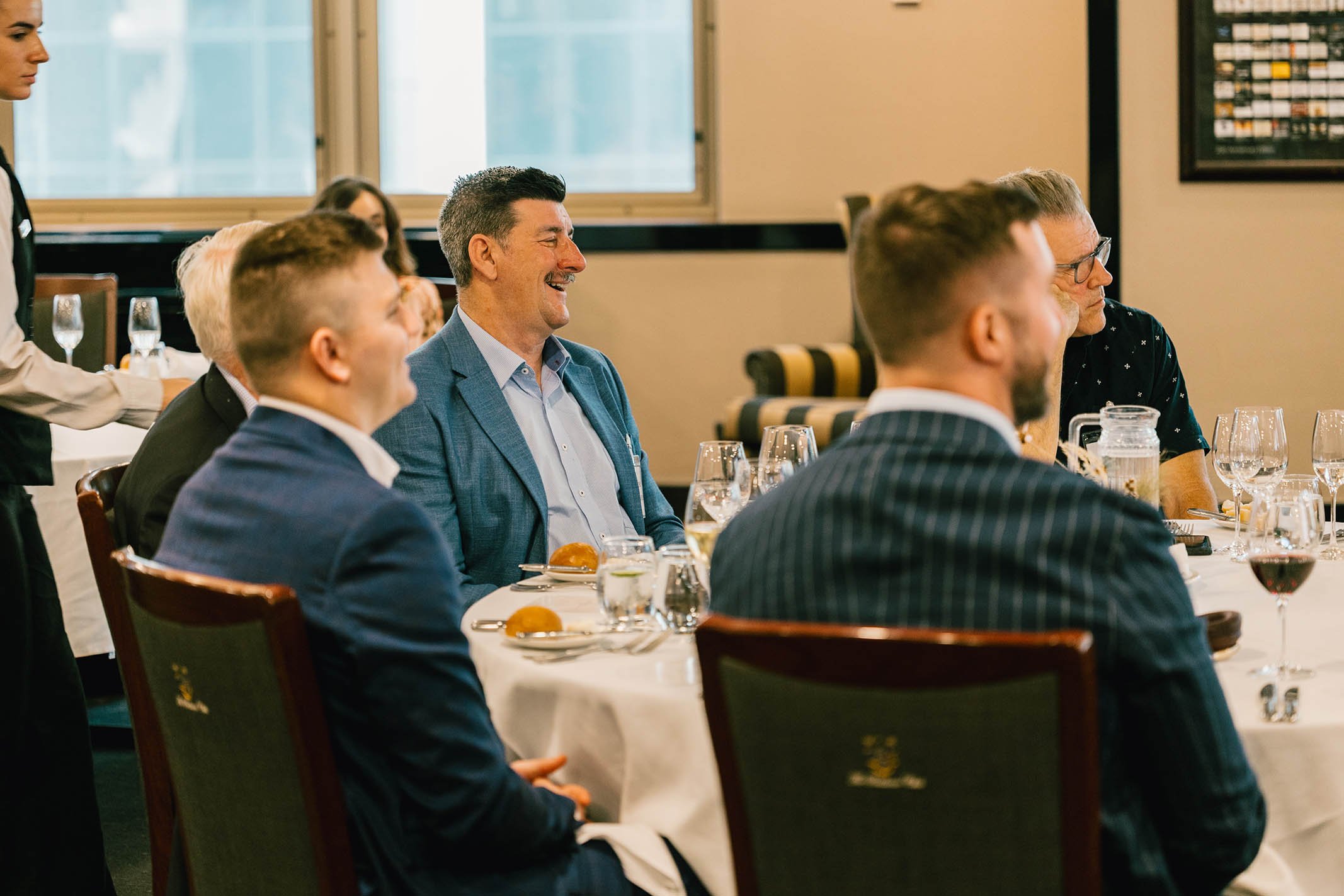 Group of men in suits and blazers sitting at a round table during a formal event, smiling and enjoying the occasion.