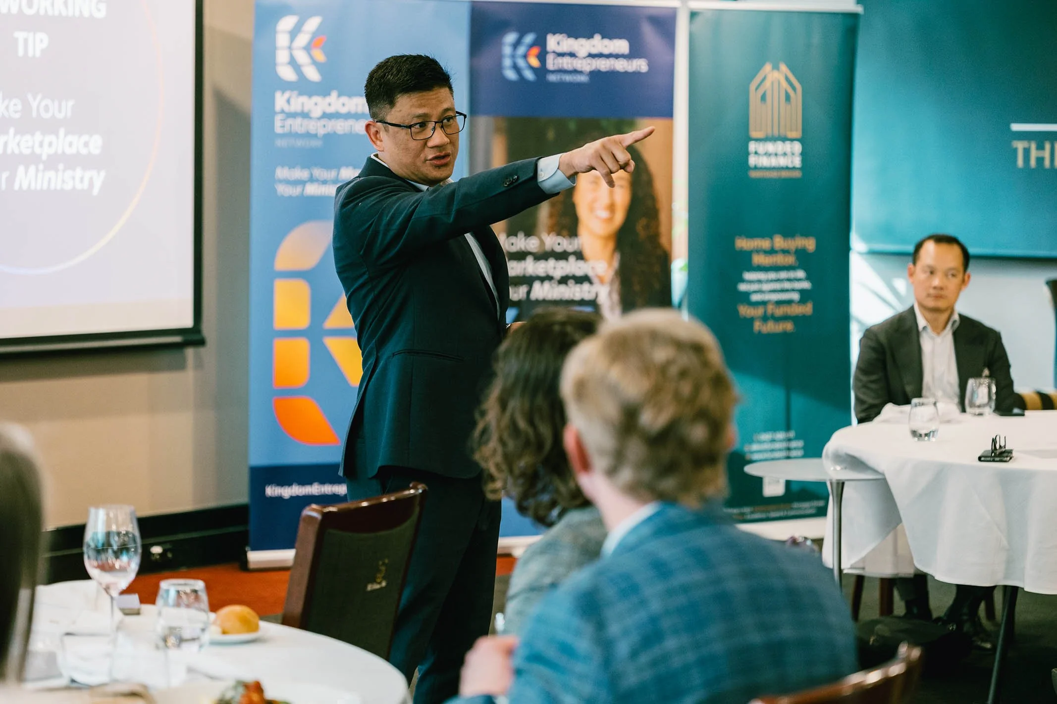A man in a dark suit and glasses is standing and speaking in front of an audience, pointing to something off-camera. There are banners in the background that read "Kingdom Entrepreneurs" and "Make Your Marketplace Your Ministry." Audience members are