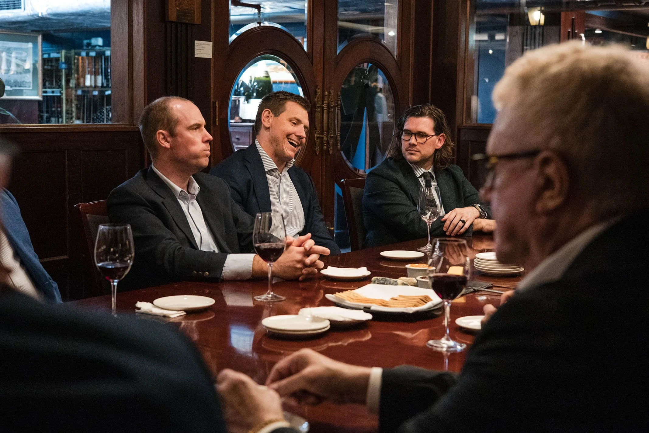 Group of men in suits sitting at a dinner table with wine glasses, plates, and snacks in a dimly lit restaurant, engaged in conversation.