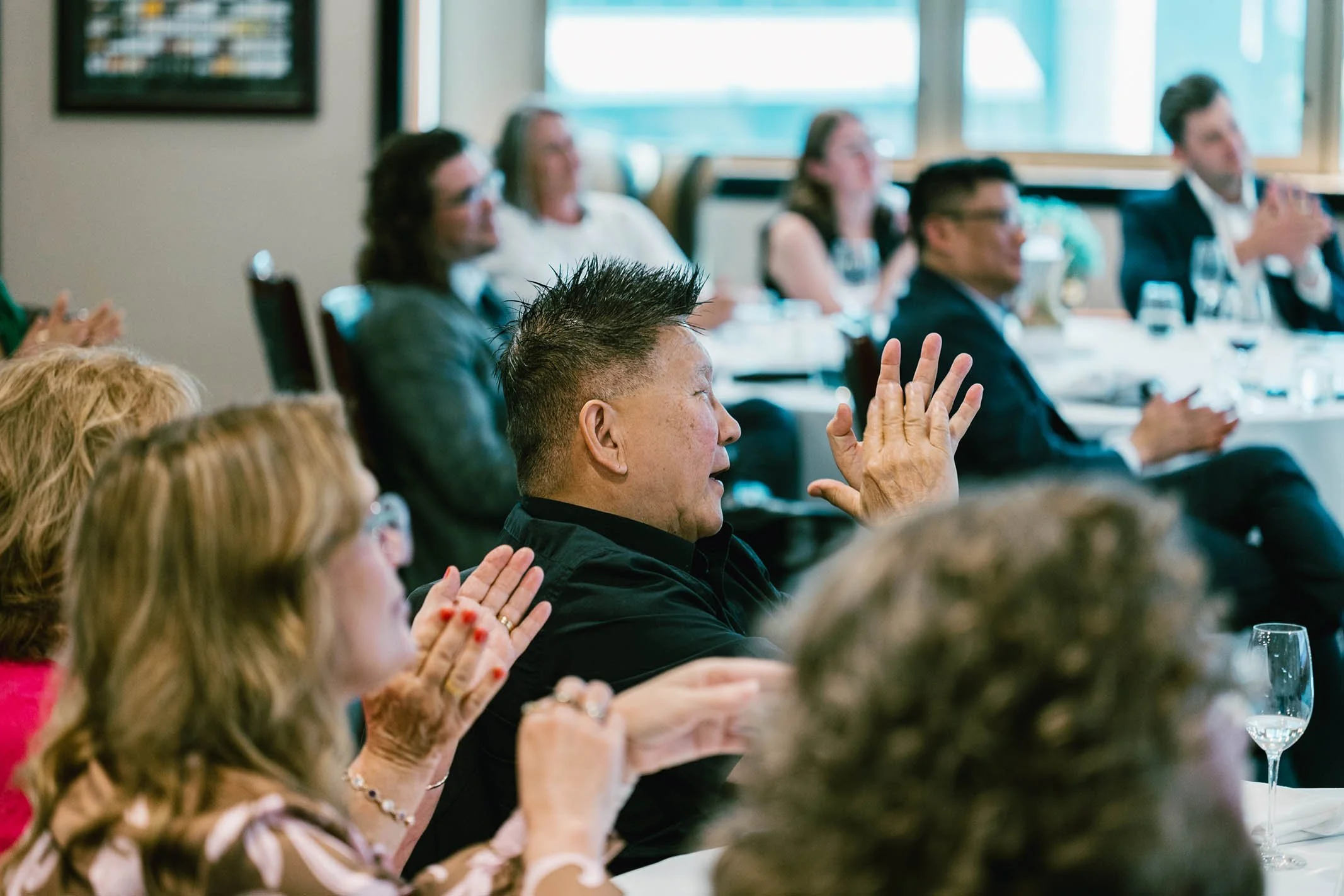 A group of people in a meeting room, clapping and listening to a speaker. The focus is on a man with spiked hair, wearing a black shirt, raising his hand.