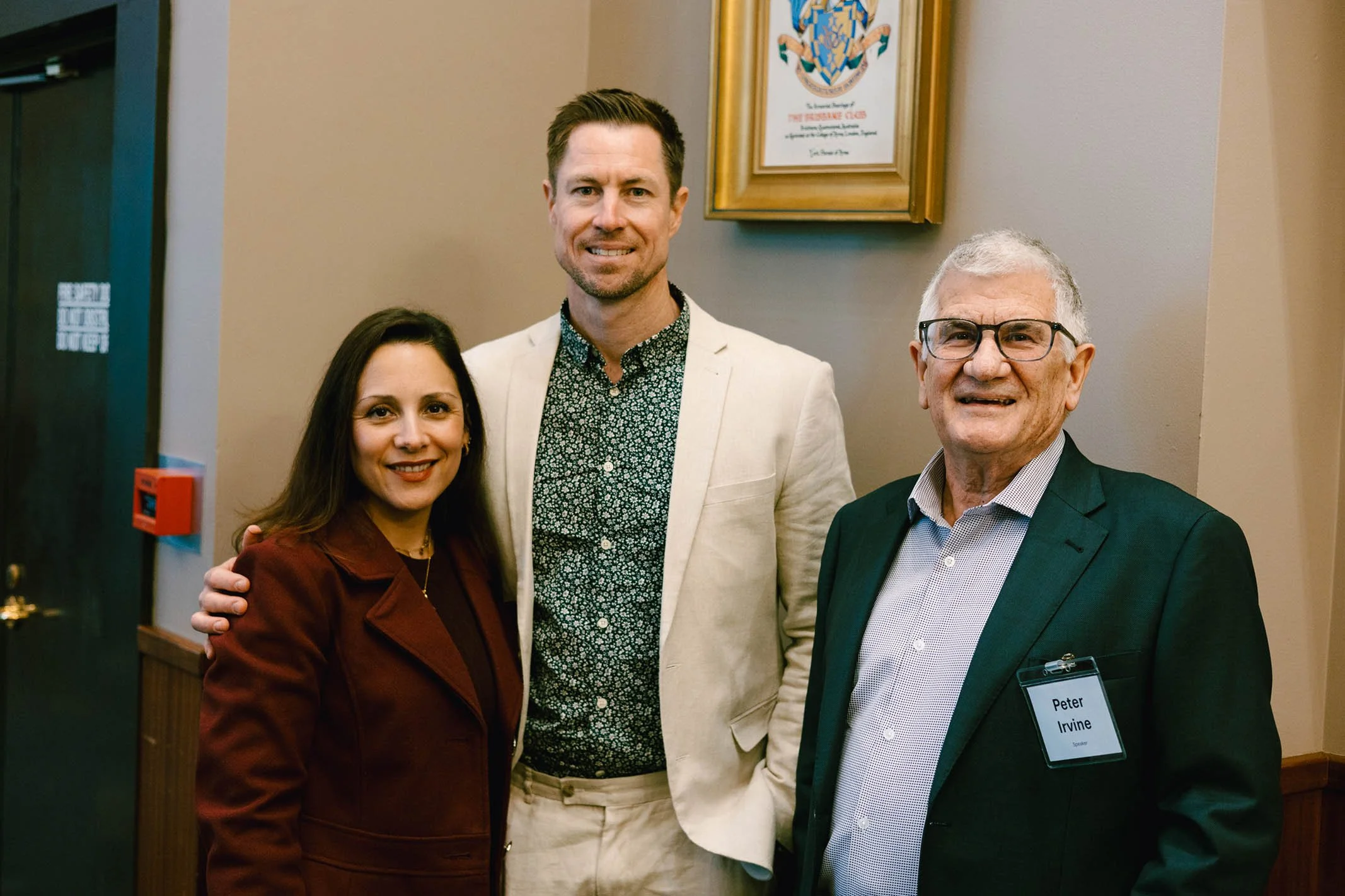 Three smiling people standing together indoors, including a woman in a maroon blazer on the left, a man in a light-colored blazer and patterned shirt in the middle, and an older man with glasses and a name tag reading 'Peter Irvine' on the right, in 