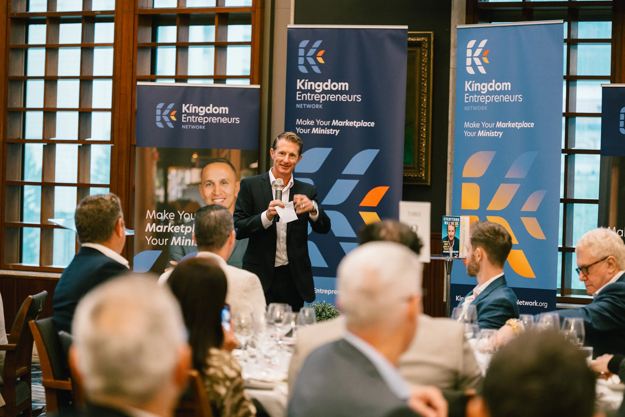 A man in a suit speaking into a microphone at a conference table surrounded by seated audience members. Banners behind him display 'Kingdom Entrepreneurs Network' and the slogan 'Make Your Marketplace Your Ministry.'