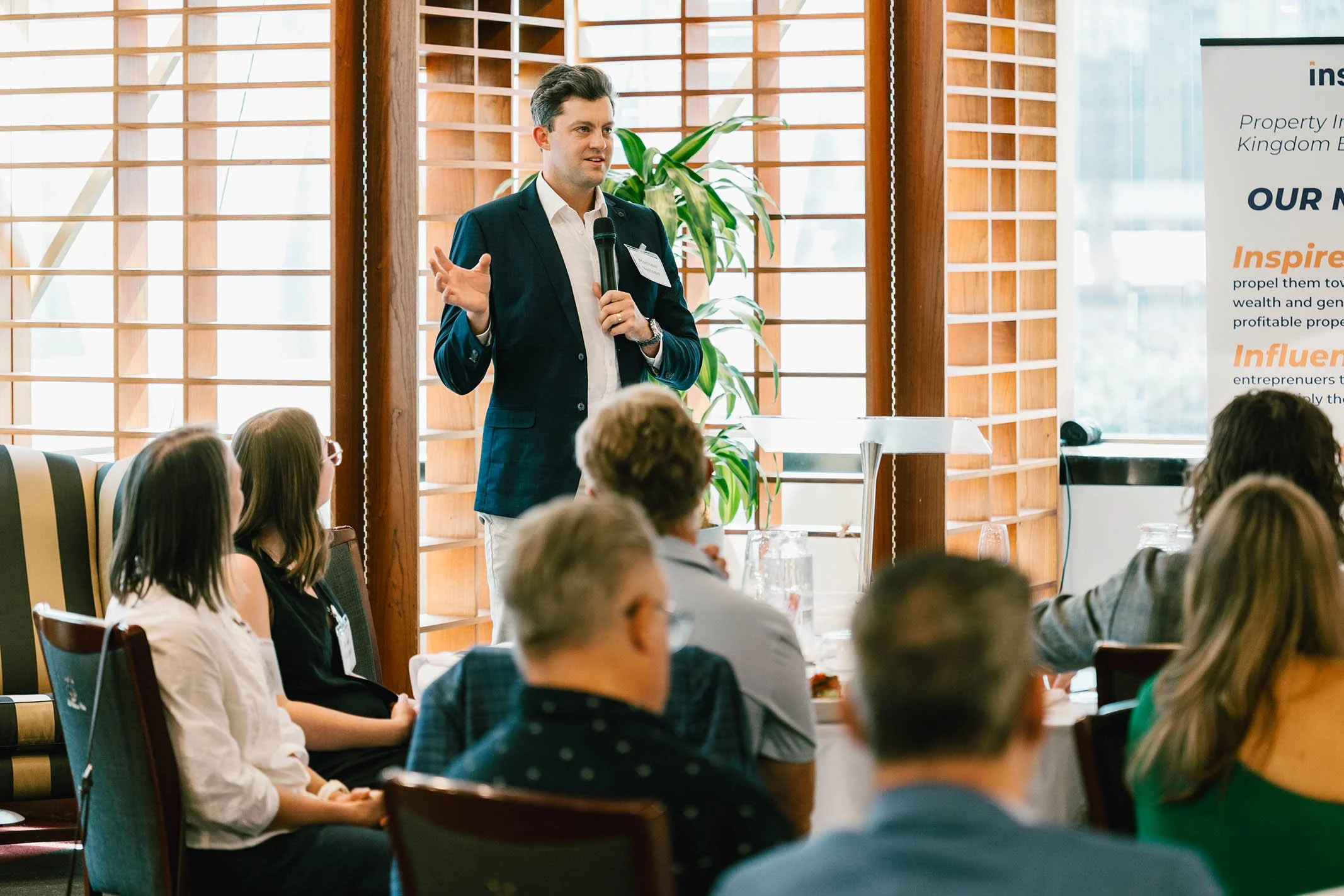 A man in a dark suit giving a presentation to a seated group of people in a room with wooden window shutters, a potted plant, and a displayed banner.