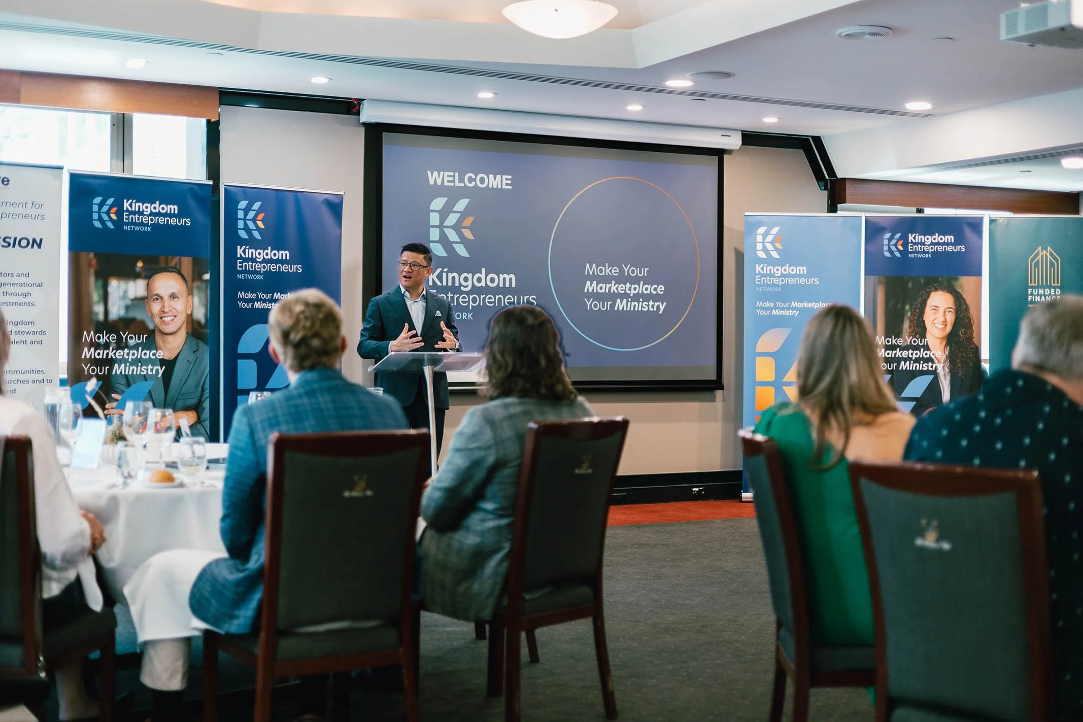 A man giving a presentation at a conference with a screen displaying 'WELCOME' and 'Make Your Marketplace Your Ministry.' Attendees are seated at round tables, some taking notes, in a conference room with banners promoting the Kingdom Entrepreneurs N