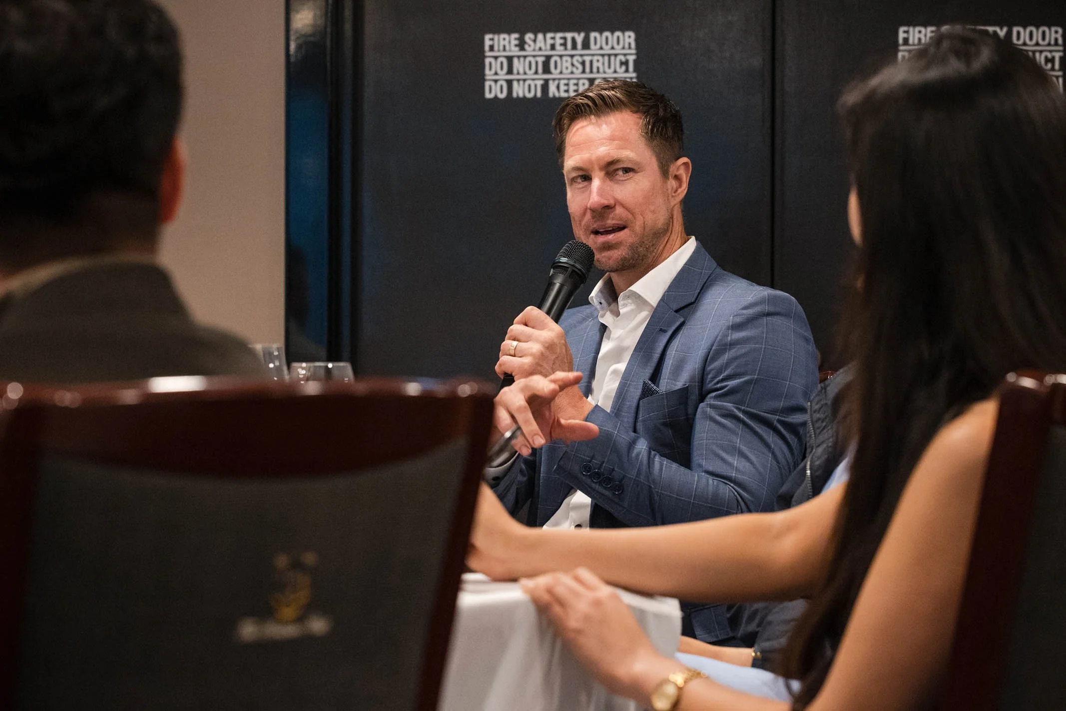 A man in a blue suit jacket speaking into a microphone at a panel discussion with an audience.