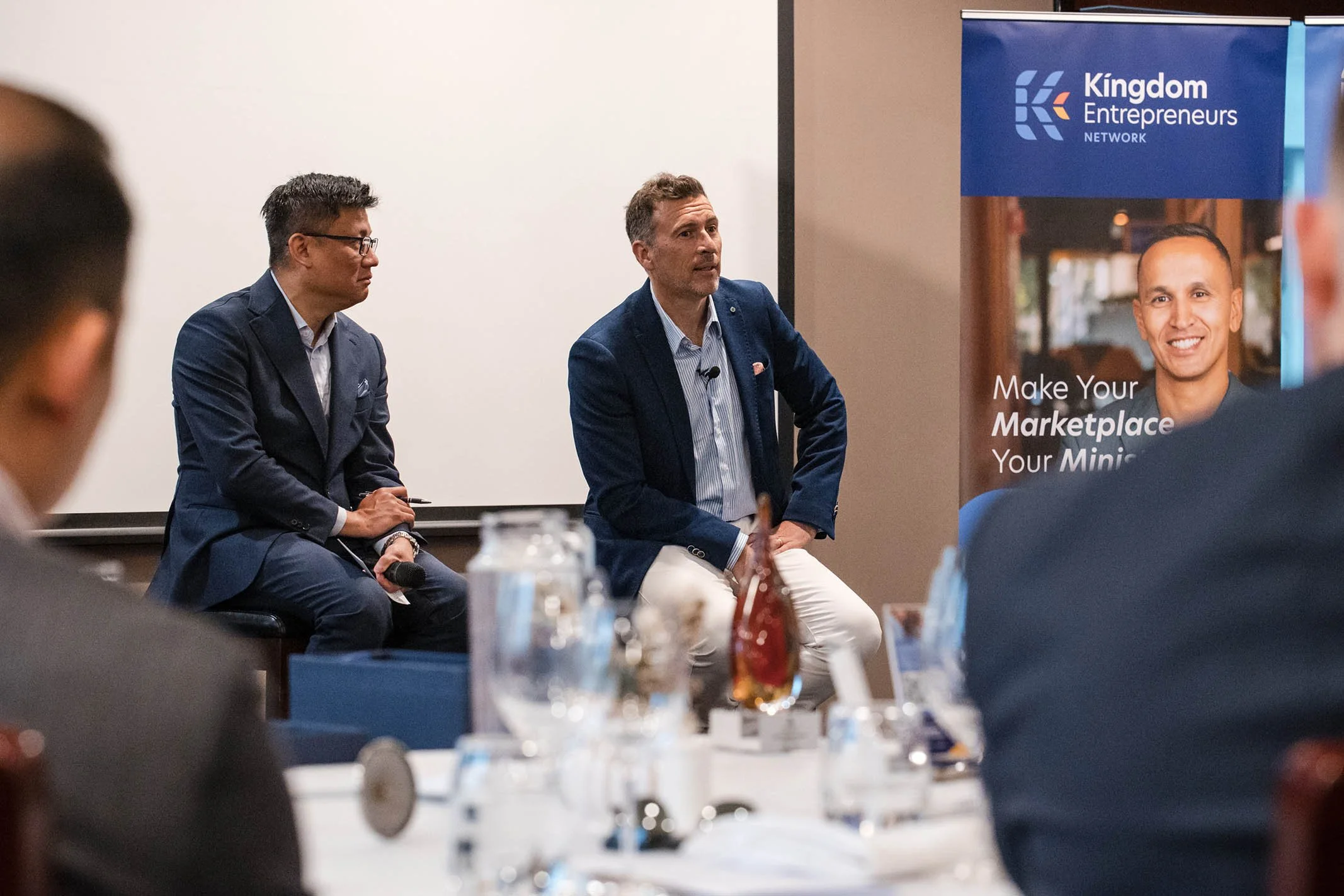 Two men sitting on stage during a professional event, with a banner in the background that reads 'Kingdom Entrepreneurs Network' and 'Make Your Marketplace Your Mini' in a conference room setting.
