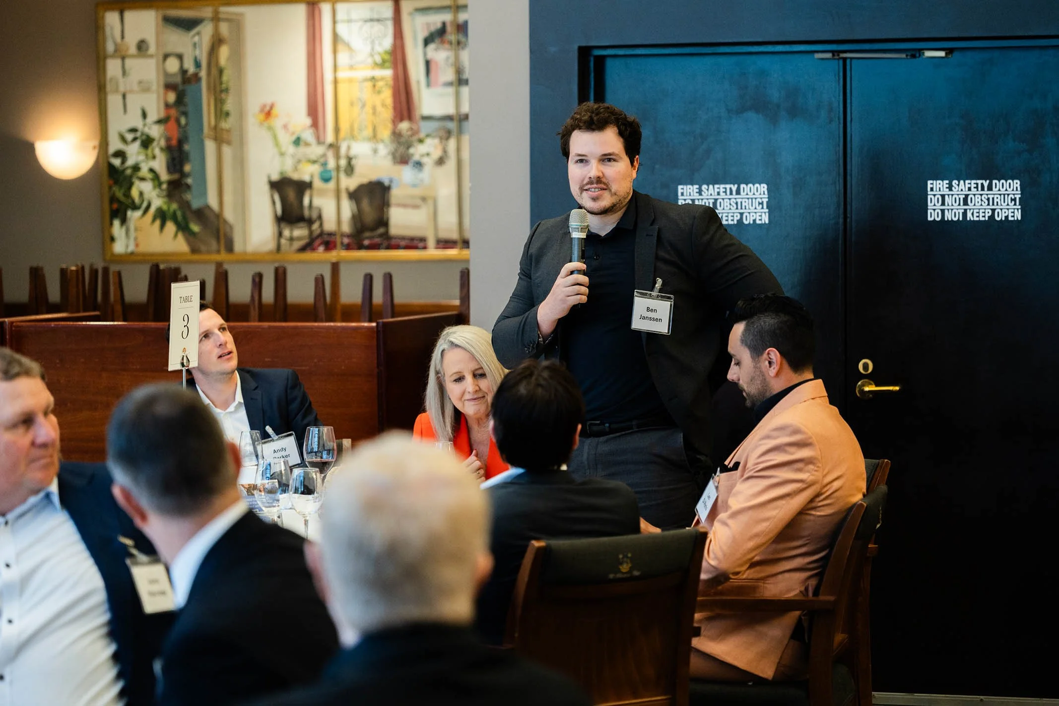Man standing and speaking into a microphone during a business meeting or conference, seated around a table with other professionally dressed individuals, some with name tags.