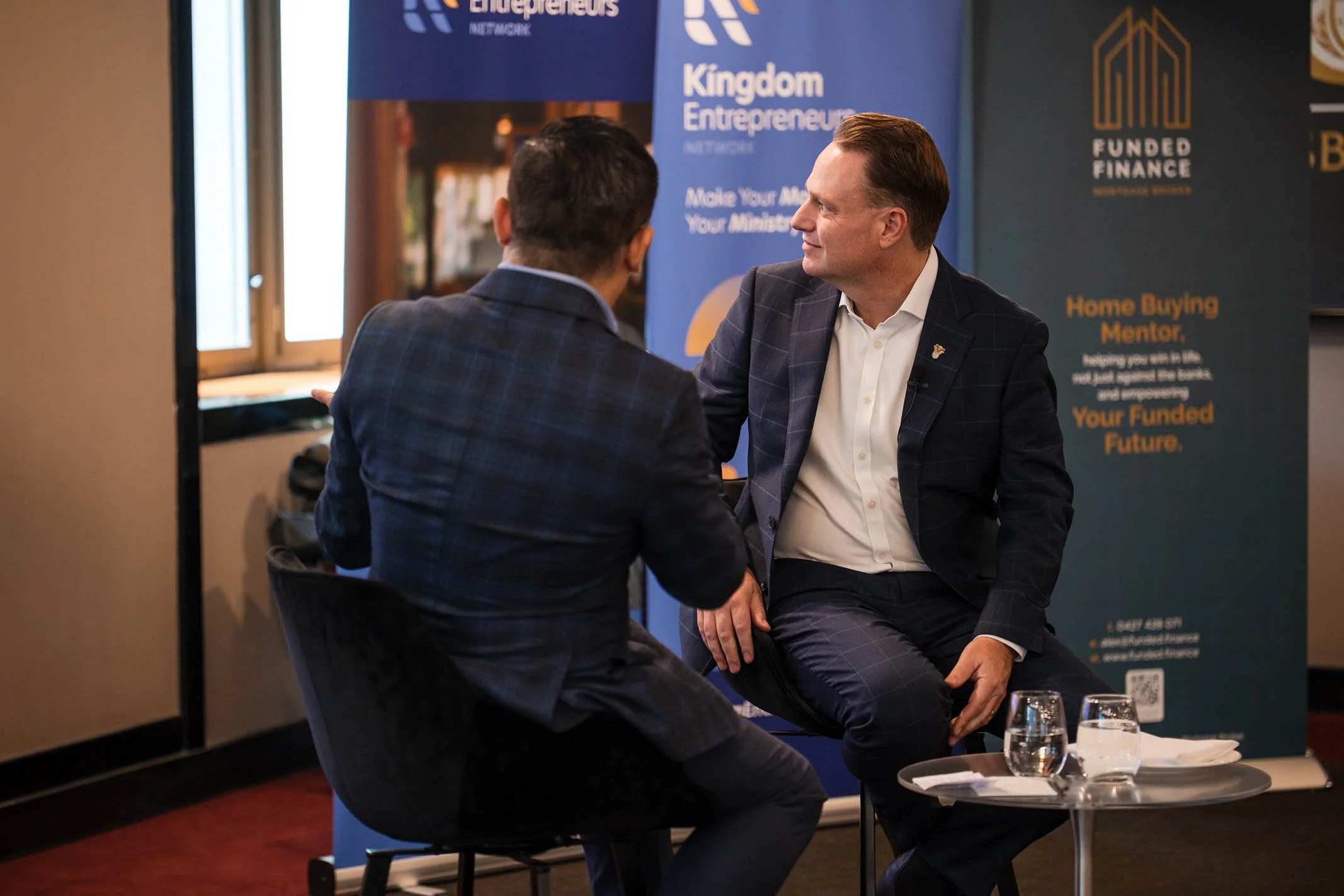 Two men in business suits engaged in a conversation at an event, seated in front of banners for Kingdom Entrepreneur Network and Funded Finance, with glasses of water and a small table between them.