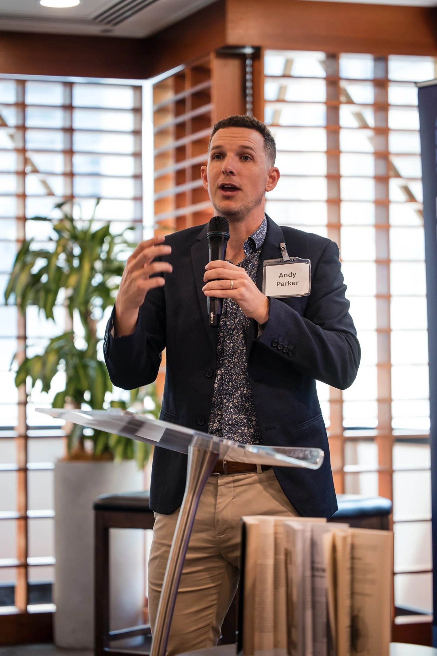 A man named Andy Parker delivering a speech at a conference, holding a microphone with one hand and gesturing with the other, in a room with wooden and glass architecture.