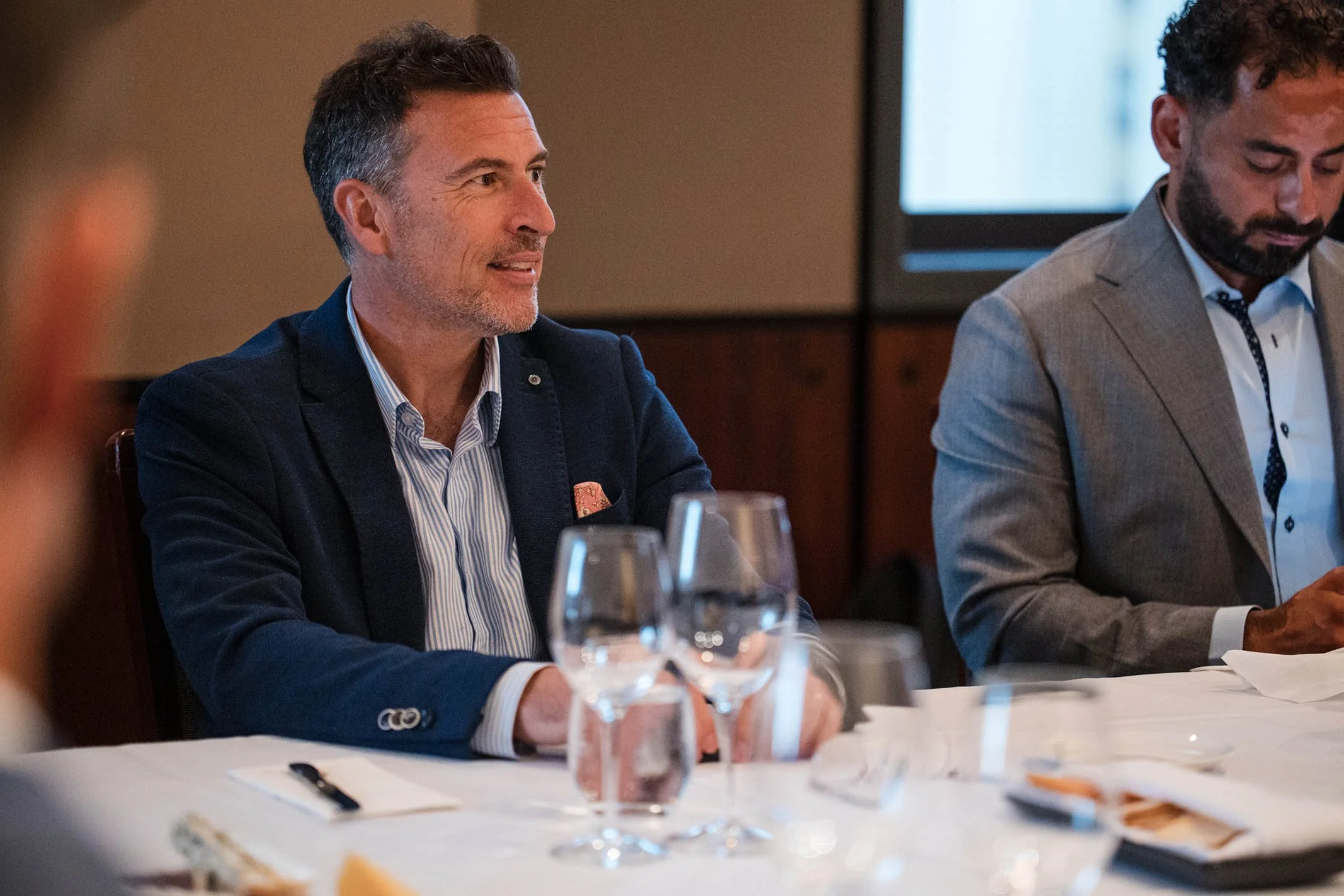 A man in a dark blue blazer and striped shirt sitting at a dinner table with glasses and plates, engaged in conversation during a formal event.