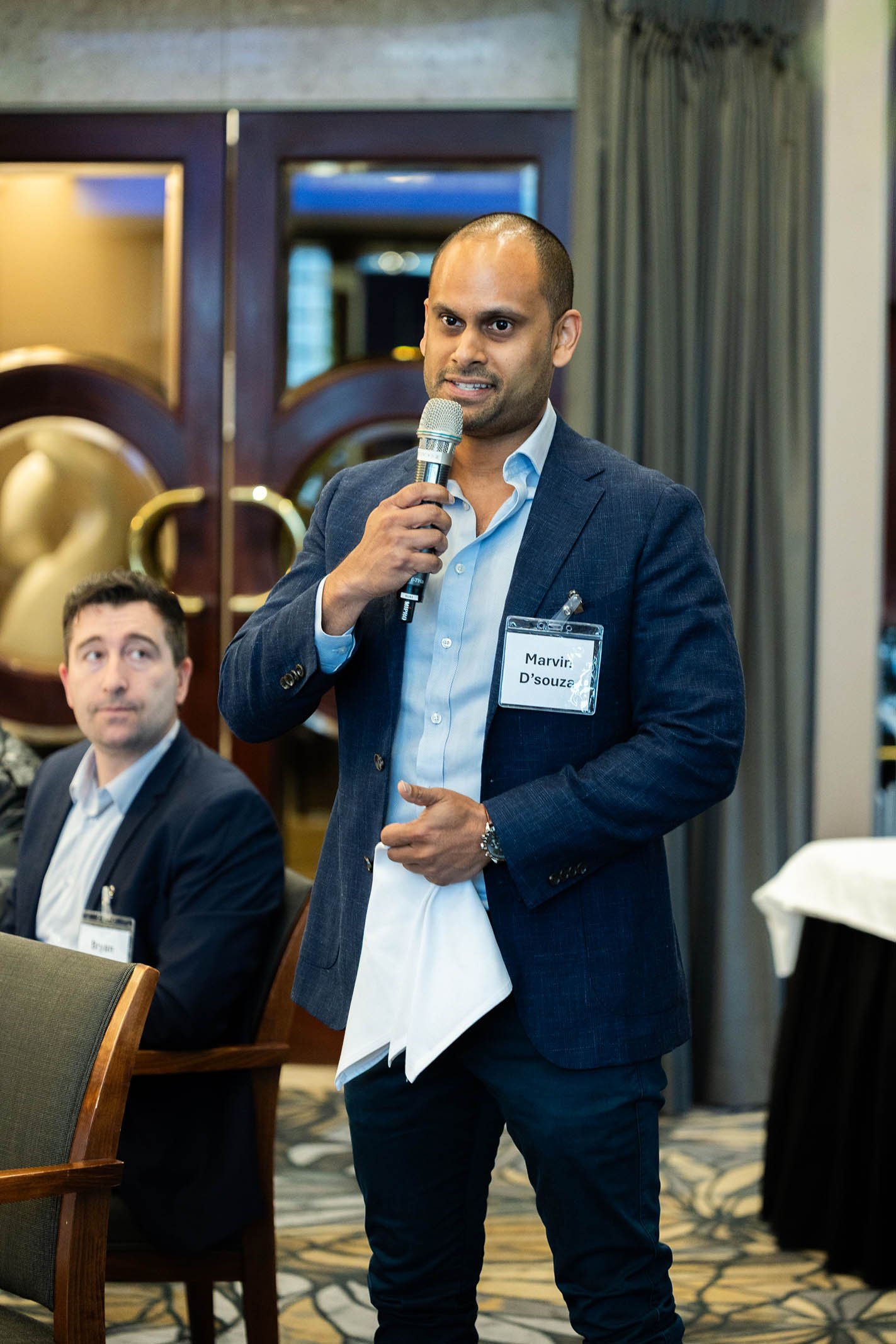 A man in a blue blazer and white shirt holding a microphone and speaking at a conference. Another man is sitting nearby, listening.