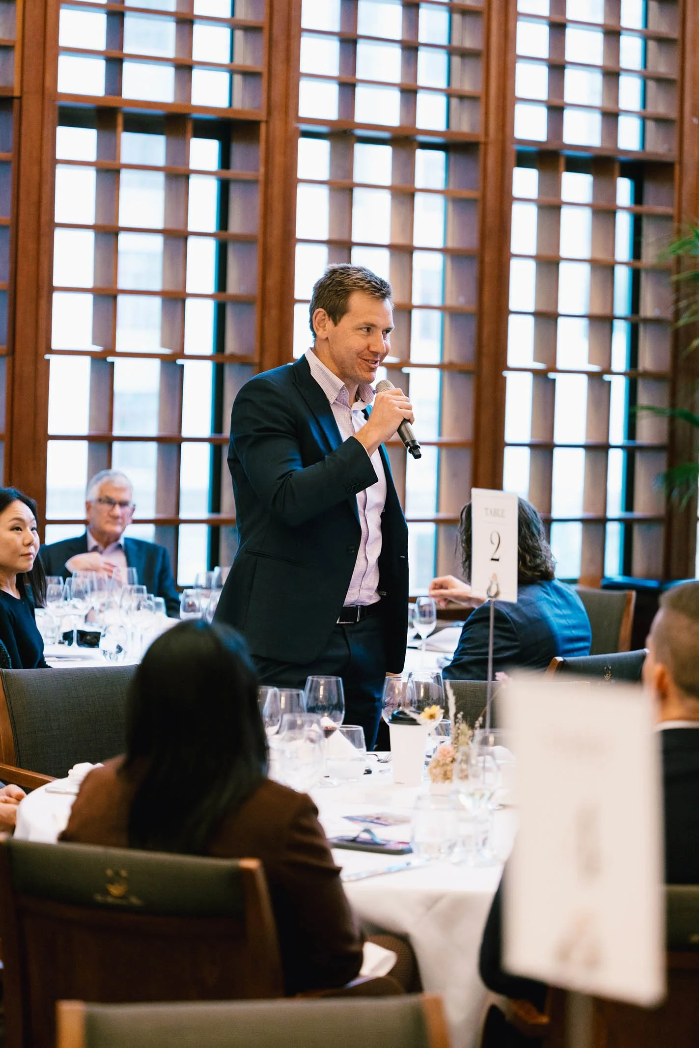A man in a dark suit is speaking into a microphone at a formal event, seated at a round table set with wine glasses and flowers, with other attendees listening.