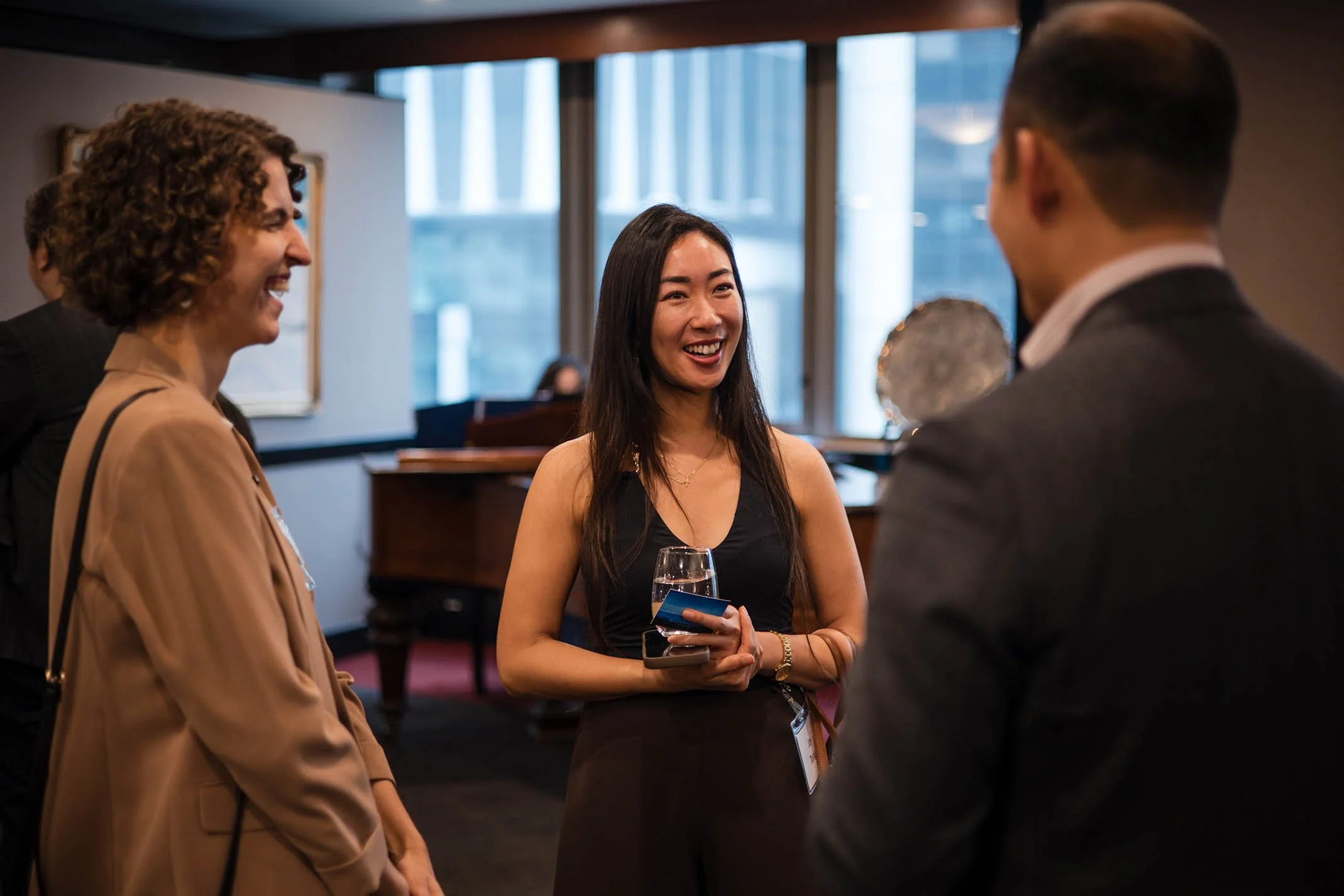 Three people engaged in conversation at a social event inside a room with large windows, a piano, and a painting on the wall.