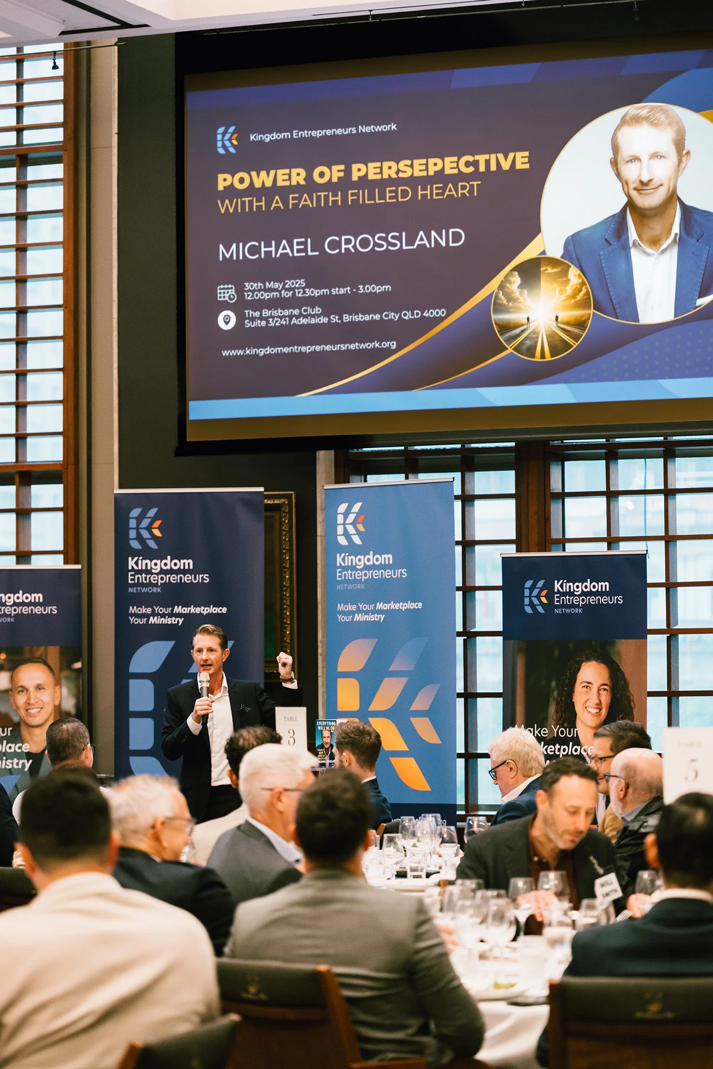 A conference room filled with people seated at tables, listening to a speaker at the front. The speaker is a man in a suit, holding a microphone and gesturing with his fist. Behind him are banners for Kingdom Entrepreneurs Network. A large screen dis