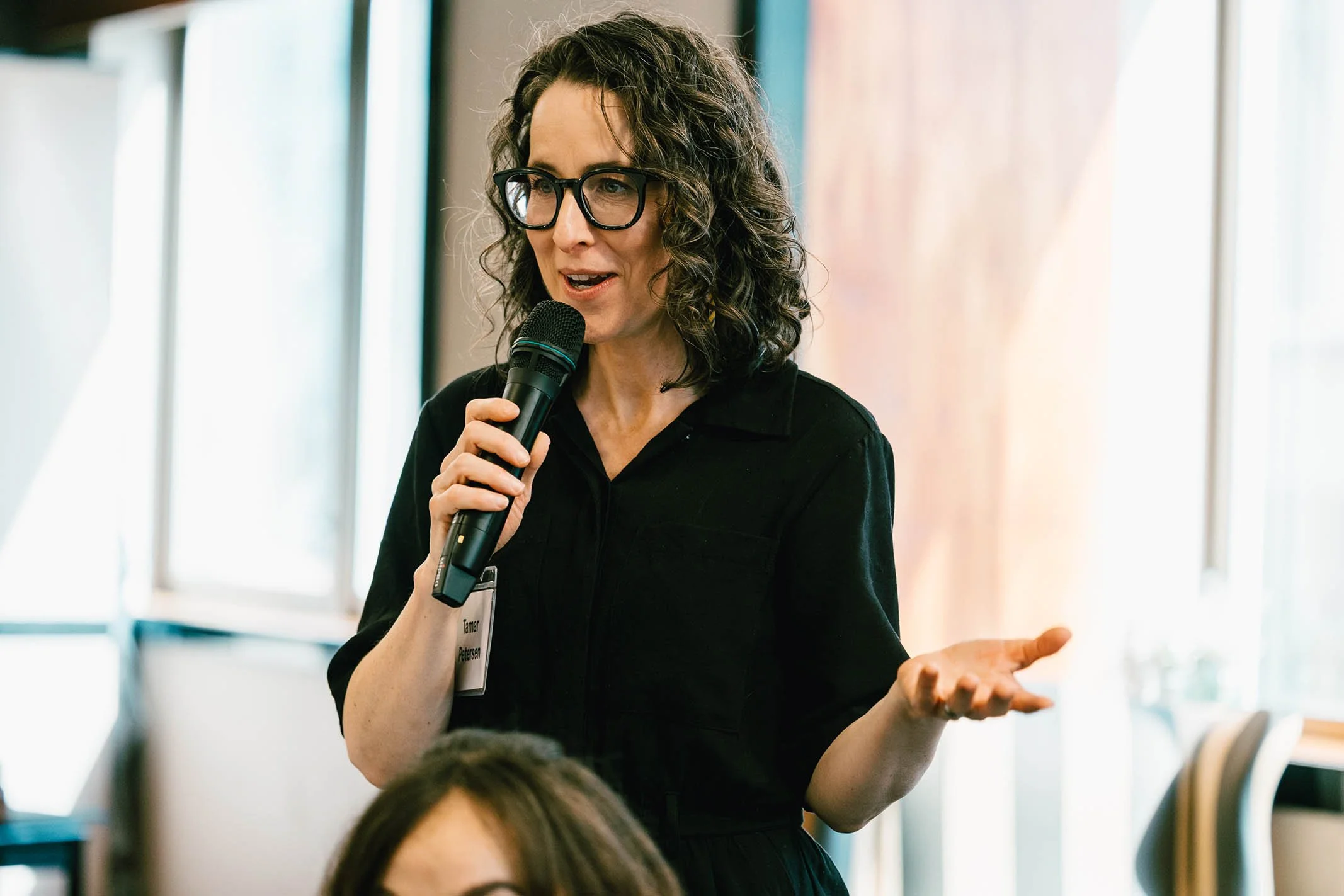 A woman with curly hair and glasses speaks into a microphone during a presentation or conference.