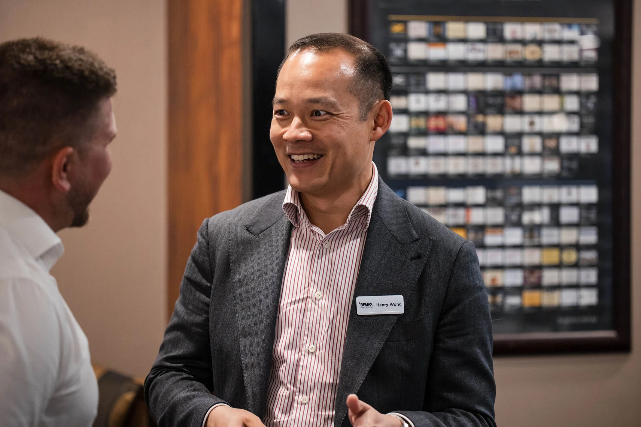Two men are conversing indoors; one is wearing a gray suit with a name tag, smiling, and the other is in a white shirt, seen from the back. There is a framed display of classified index cards in the background.