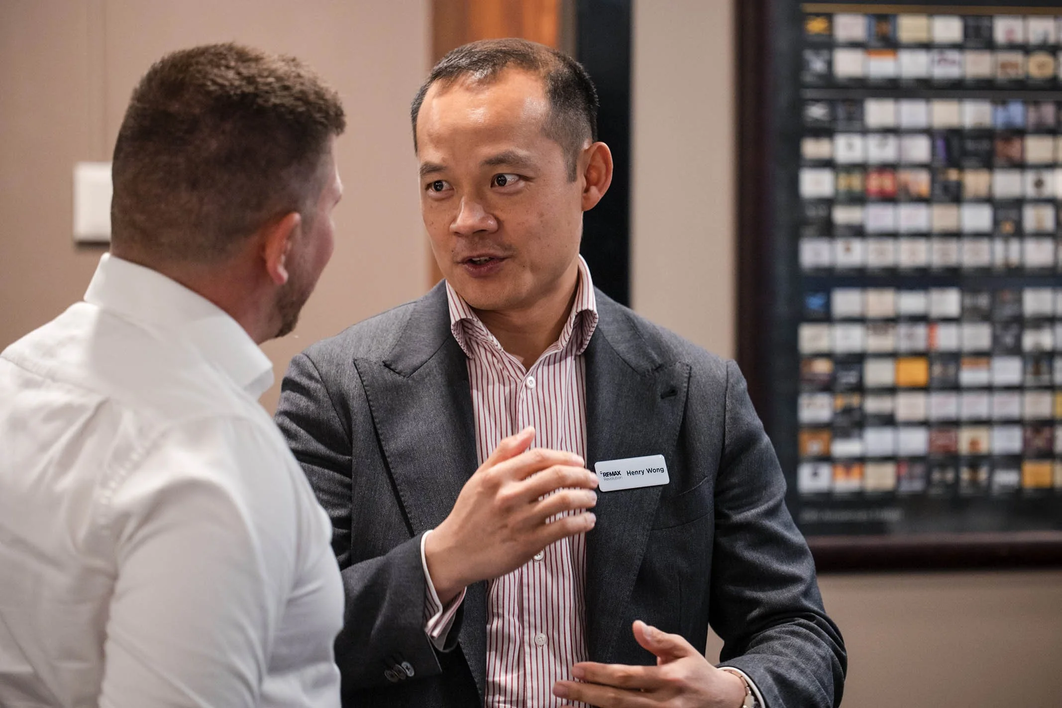 Two men are engaged in conversation in an indoor setting, with one wearing a white shirt and the other in a grey blazer with a name tag that reads "Henry Wong," in front of a framed collection of small rectangular items on the wall.