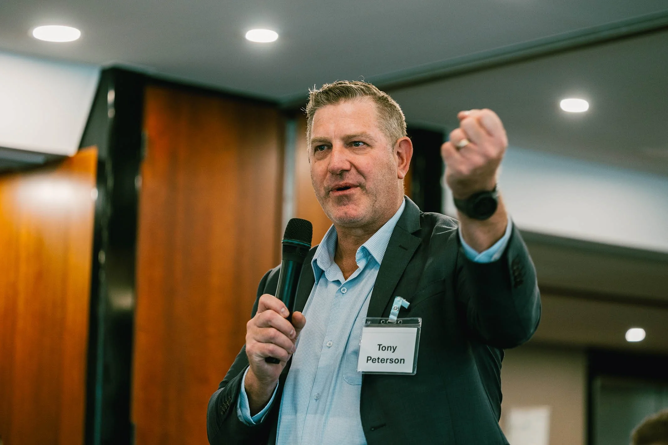 A man in a suit holding a microphone and making a fist gesture while speaking at an indoor event, wearing a name badge that reads 'Tony Peterson'.