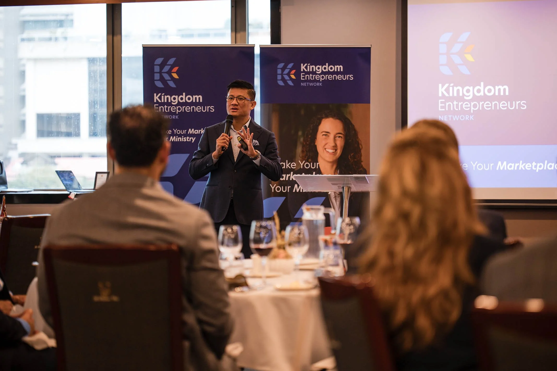 A man in a suit is giving a presentation at a conference, holding a microphone and gesturing with his right hand. Behind him are banners that read 'Kingdom Entrepreneurs Network,' and a large screen displays the same text and slogan. The audience is 