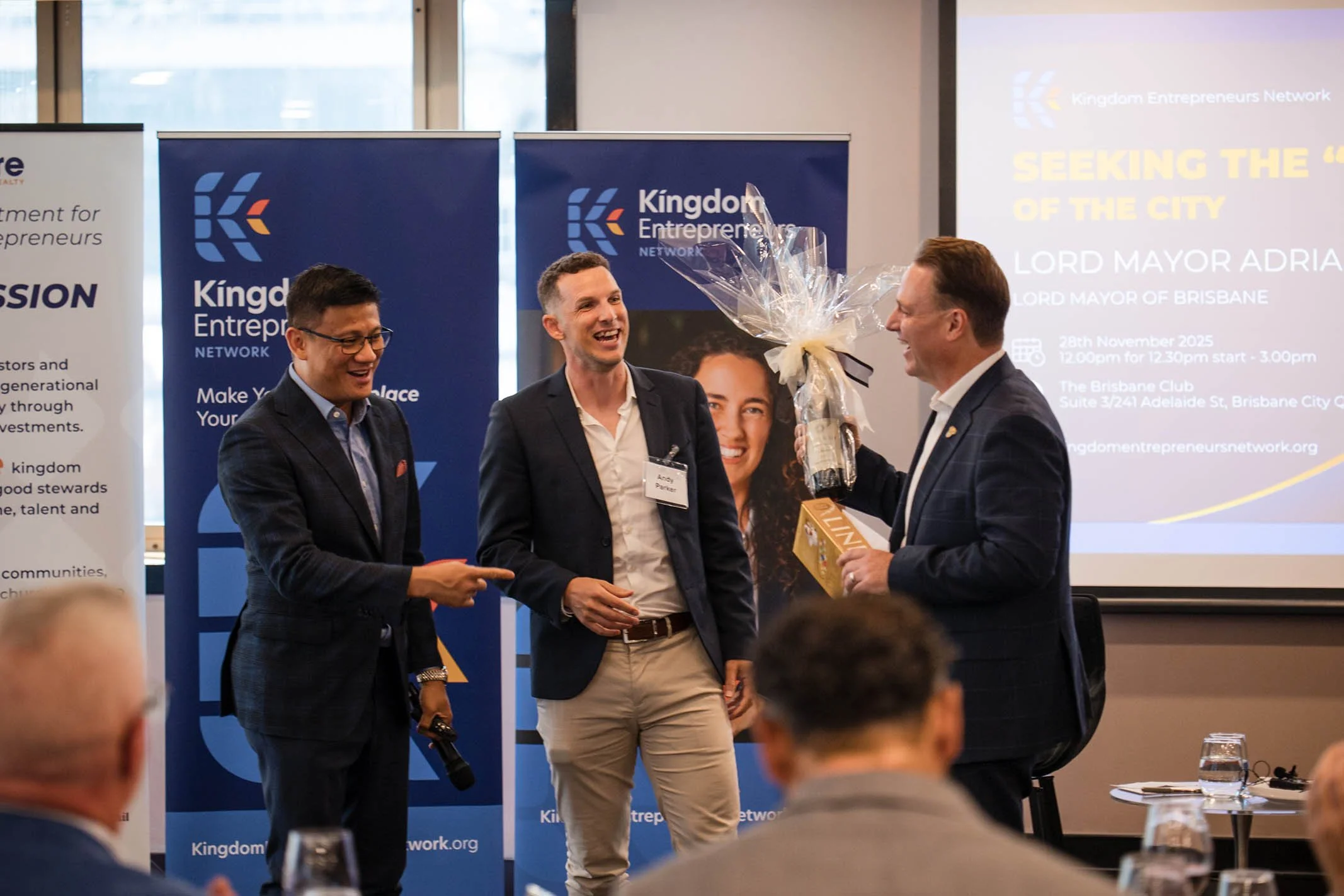 Three men at a professional event with banners in the background that read 'Kingdom Entrepreneurs Network' and a projector screen with event details, one man presents a bouquet of flowers to another man.