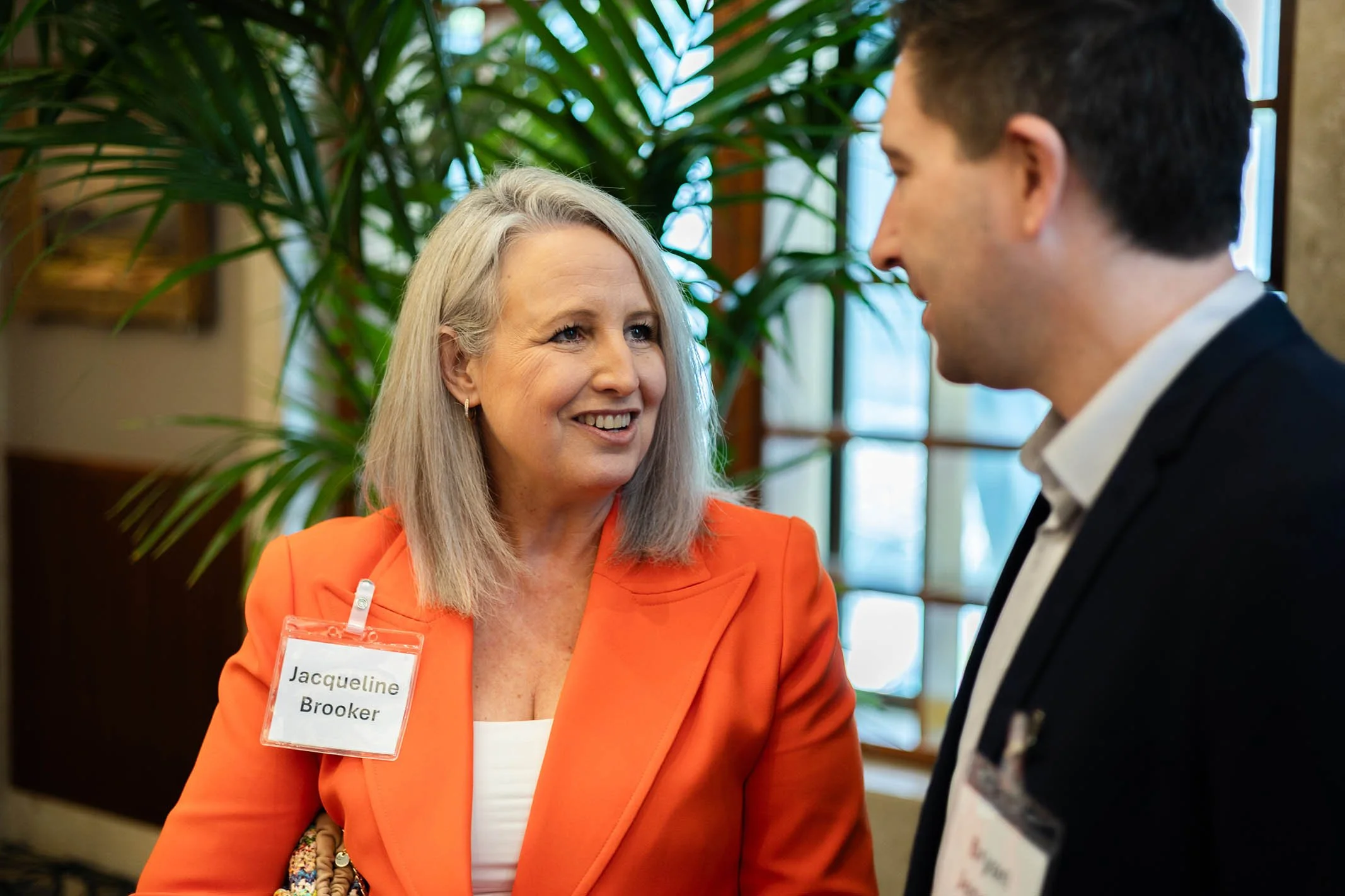 A woman with blonde hair wearing an orange blazer and a name tag that says 'Jacqueline Brooker' is smiling and talking to a man wearing a dark suit and white shirt, in an indoor setting with green plants and large windows.