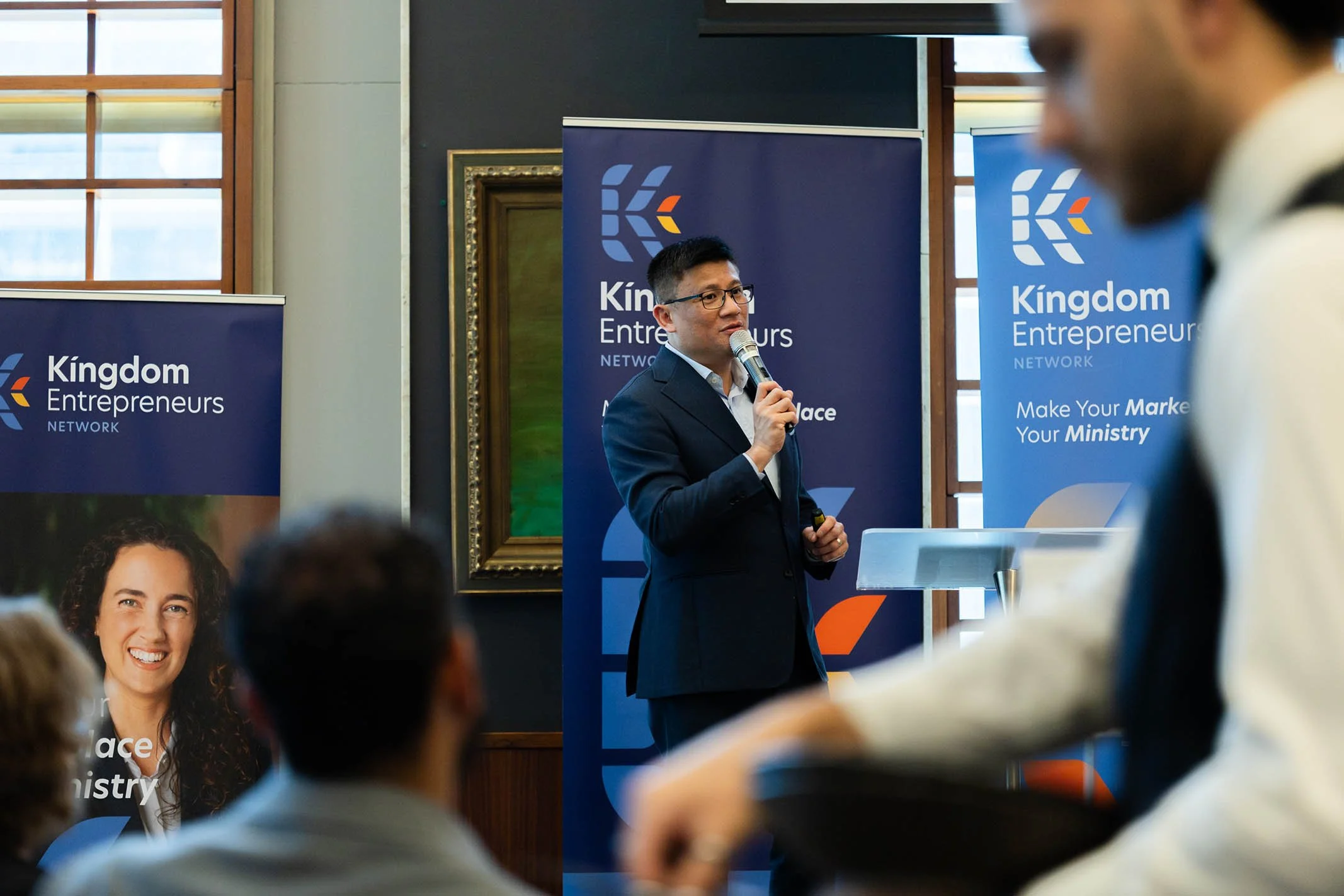 A man in a suit speaking into a microphone at a Kingdom Entrepreneurs Network event, with banners and attendees in the background.