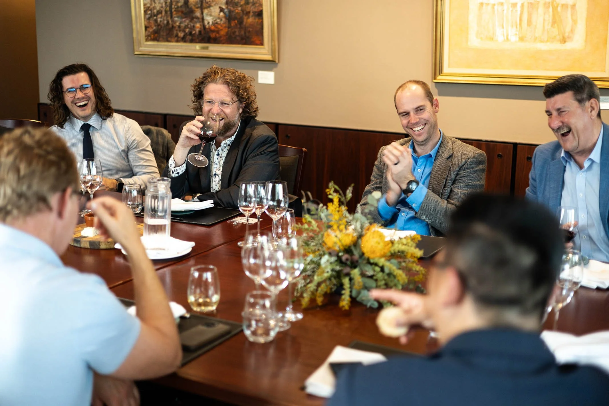 A group of seven people sitting around a wooden table, laughing and enjoying drinks, with a centerpiece of yellow and green flowers on the table.