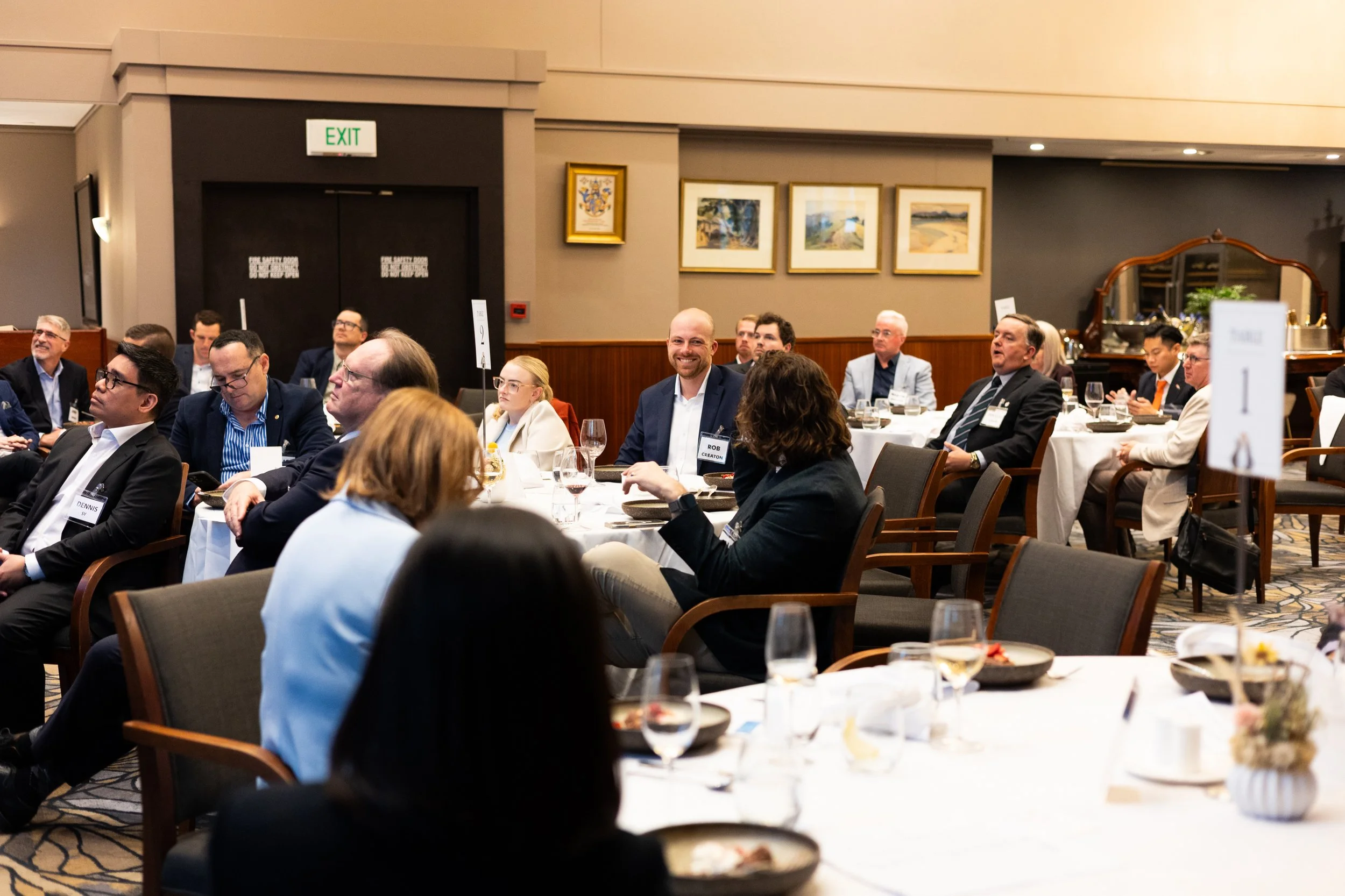 People attending a conference or formal event seated at round tables in a well-lit room, some engaging and smiling, with side tables, wine glasses, and floral centerpieces.