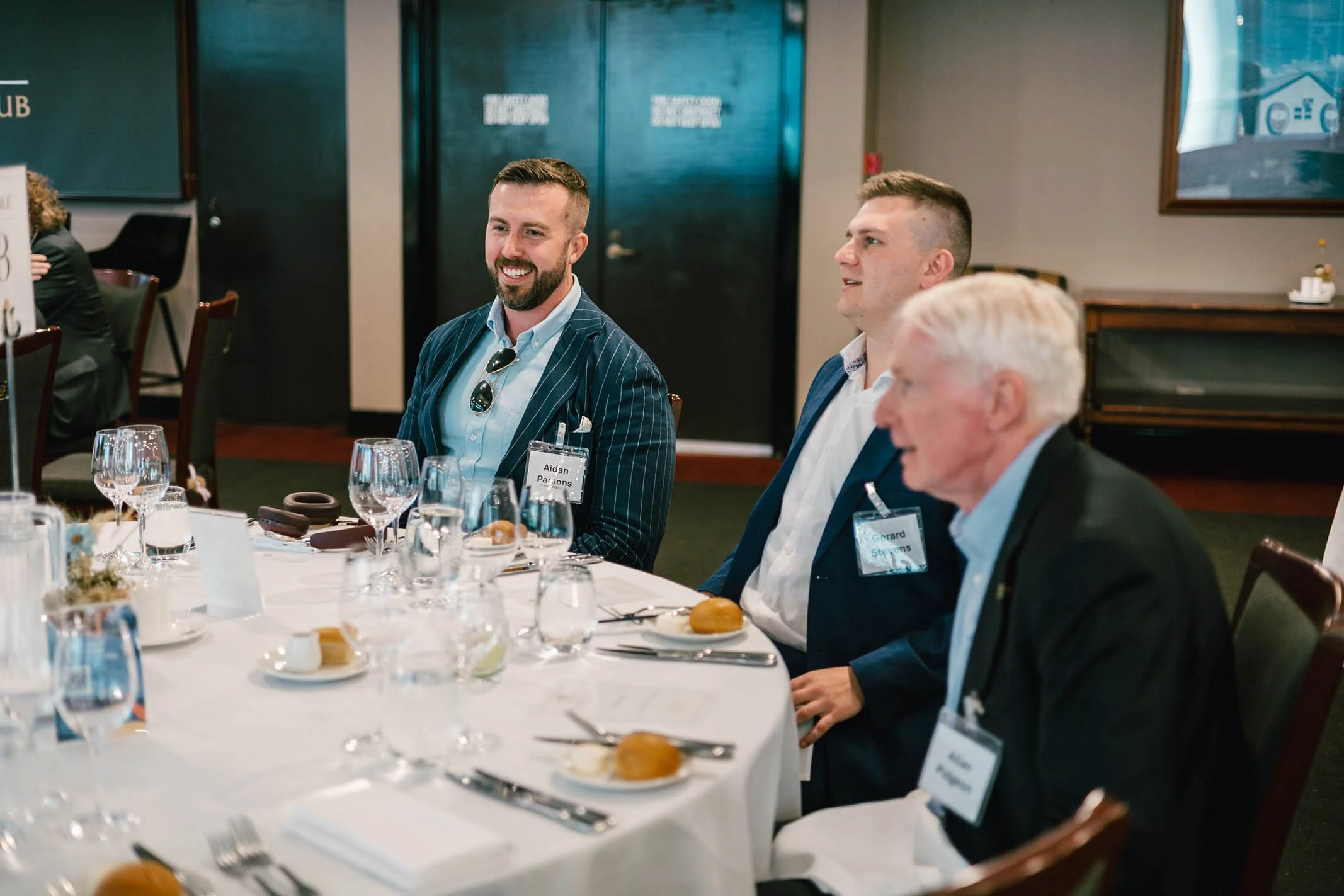 Three men in suits sitting at a round table with white tablecloth, glassware, plates with bread, and silverware in a formal dining setting, smiling and engaging in conversation.