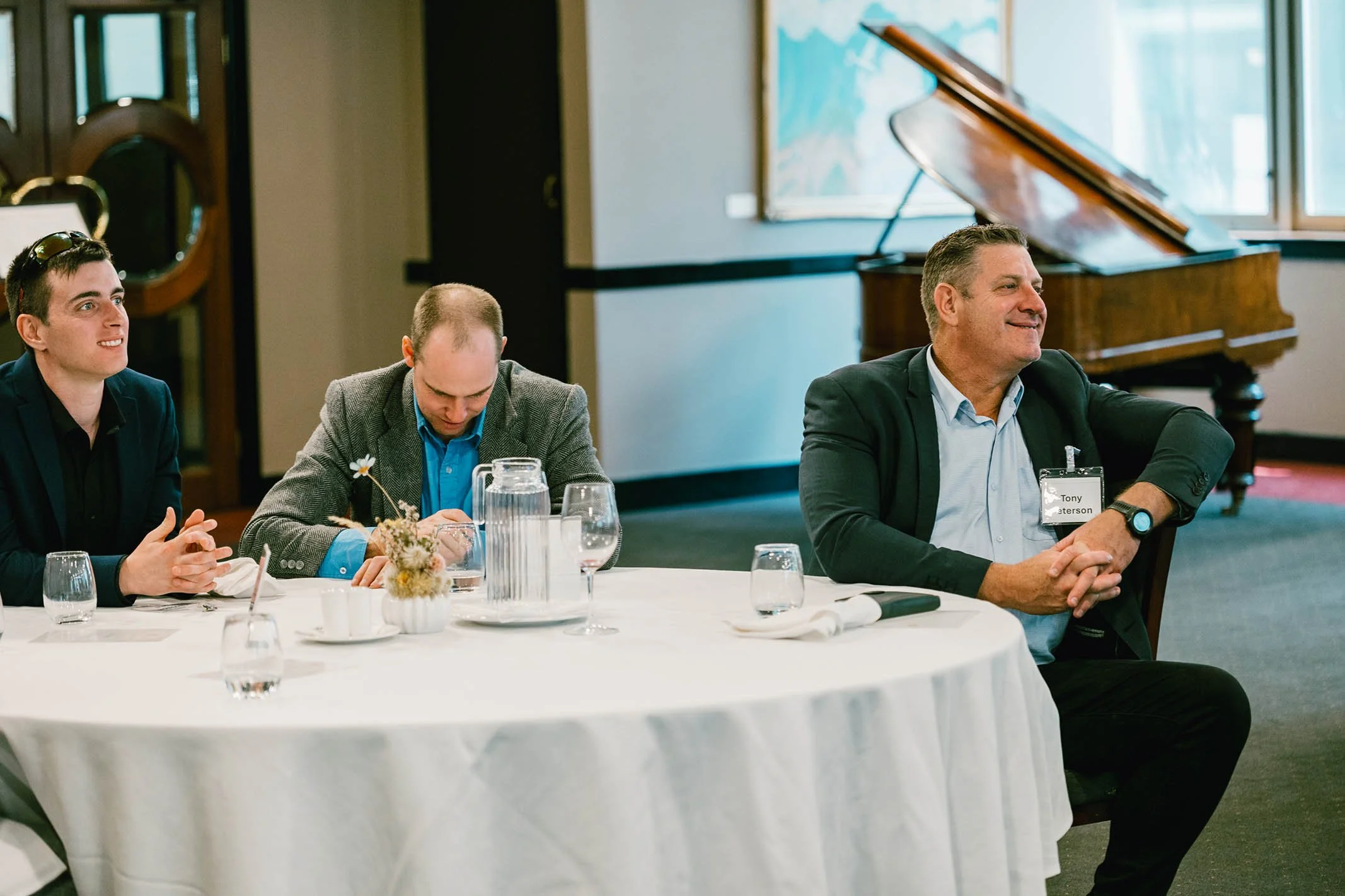 Three men sitting at a round conference table during a meeting or event. The man on the right is smiling, wearing a dark blazer, light blue shirt, and a name tag. The middle man is looking down at his phone, dressed in a blue shirt and gray blazer. T