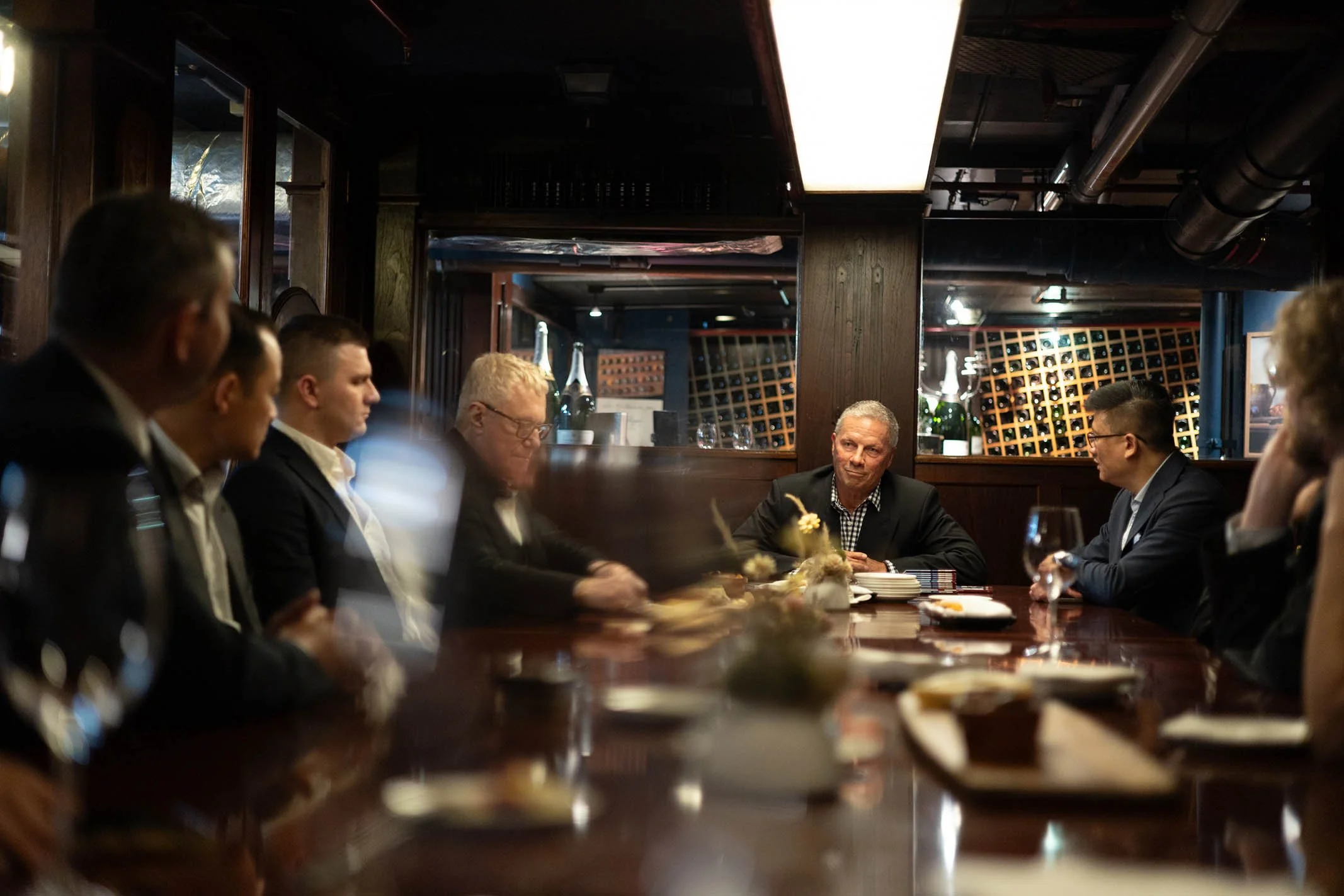 A group of people, mostly men in formal attire, sitting around a long wooden table in a restaurant or wine cellar, engaging in conversation. The setting is dimly lit with wine bottles and a wine rack visible in the background.