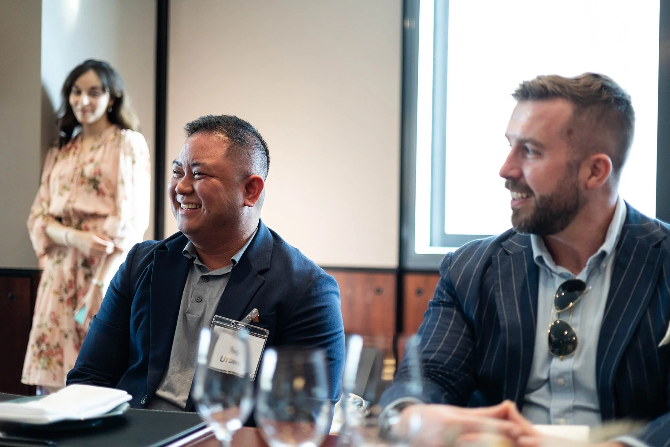 Two men sitting at a table, smiling and engaging, with a woman standing in the background, in a conference or meeting room.