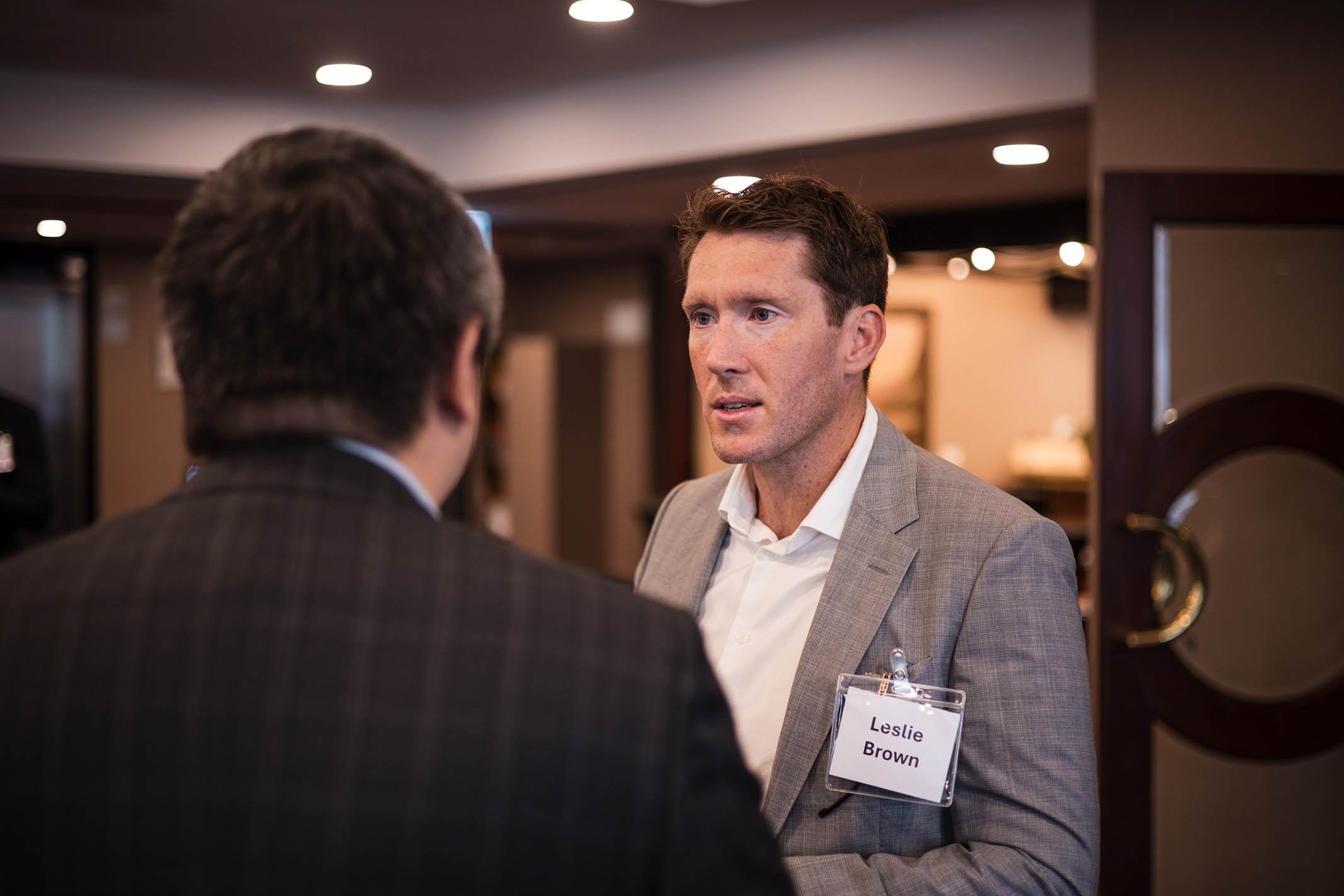 A man with a name tag reading 'Leslie Brown' speaking to another person at a professional event or conference in a well-lit indoor setting.