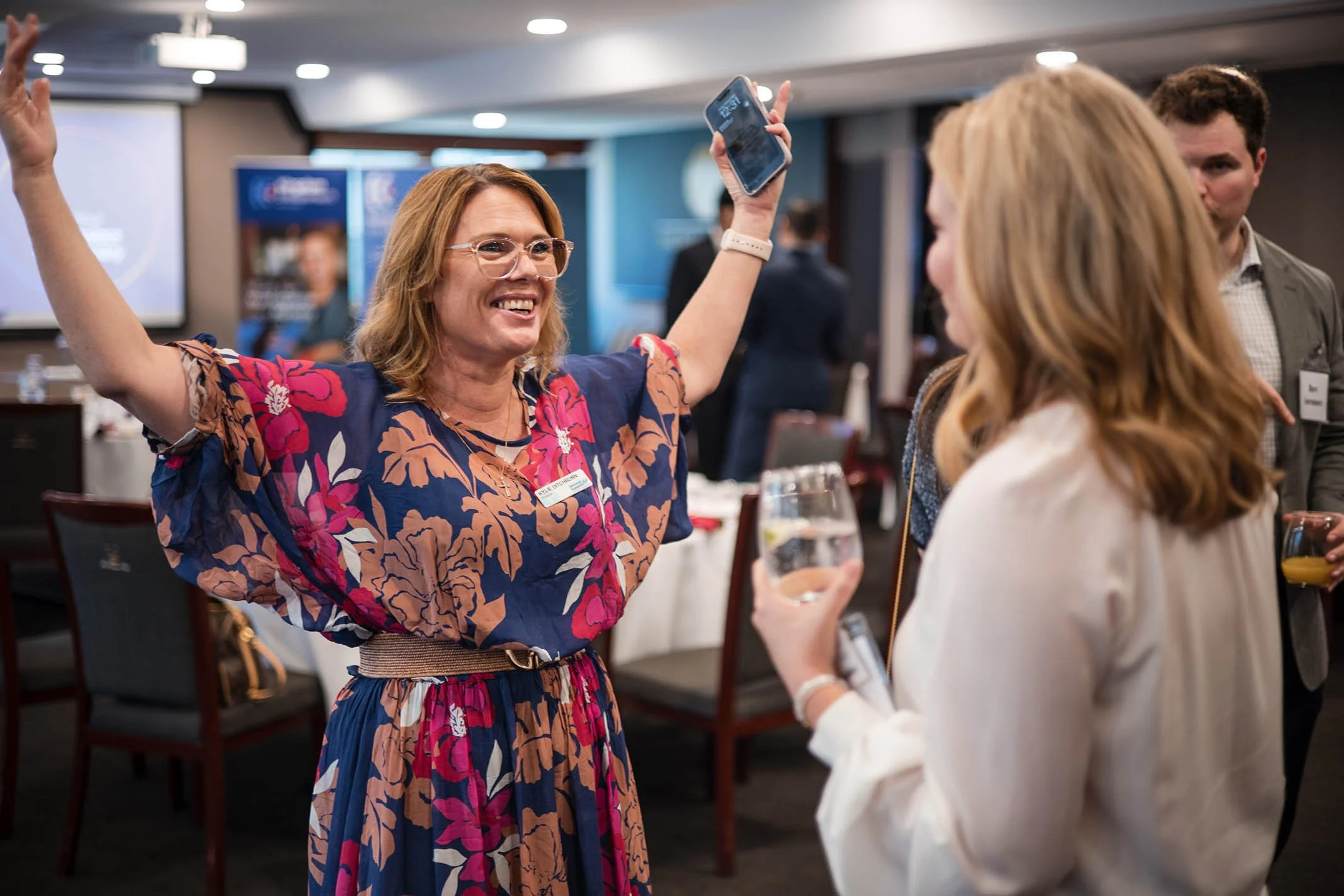 Two women are celebrating at a professional event, one with arms raised and smiling, the other holding a glass of water, with people and a presentation screen in the background.