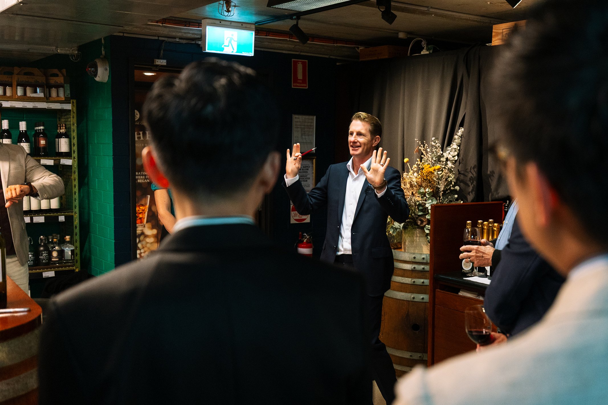 A man in a dark suit is giving a speech or presentation to an audience at an indoor event, with wine glasses in hand. The setting appears to be a cozy, decorated space with shelves of bottles and a floral arrangement in the background.