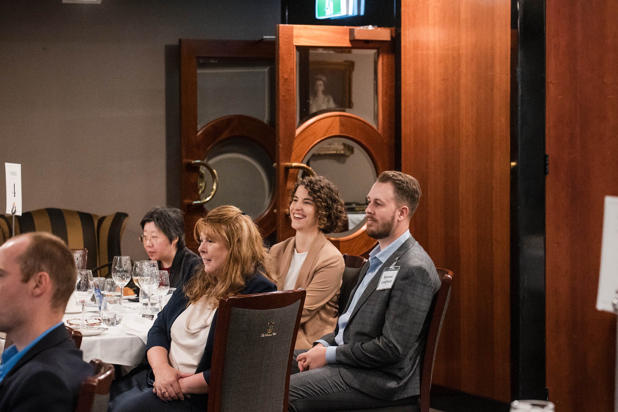 People seated at a formal dinner table, smiling and listening to a speaker or presentation, in a restaurant or event space with wooden decor and glasses of water and wine on the table.