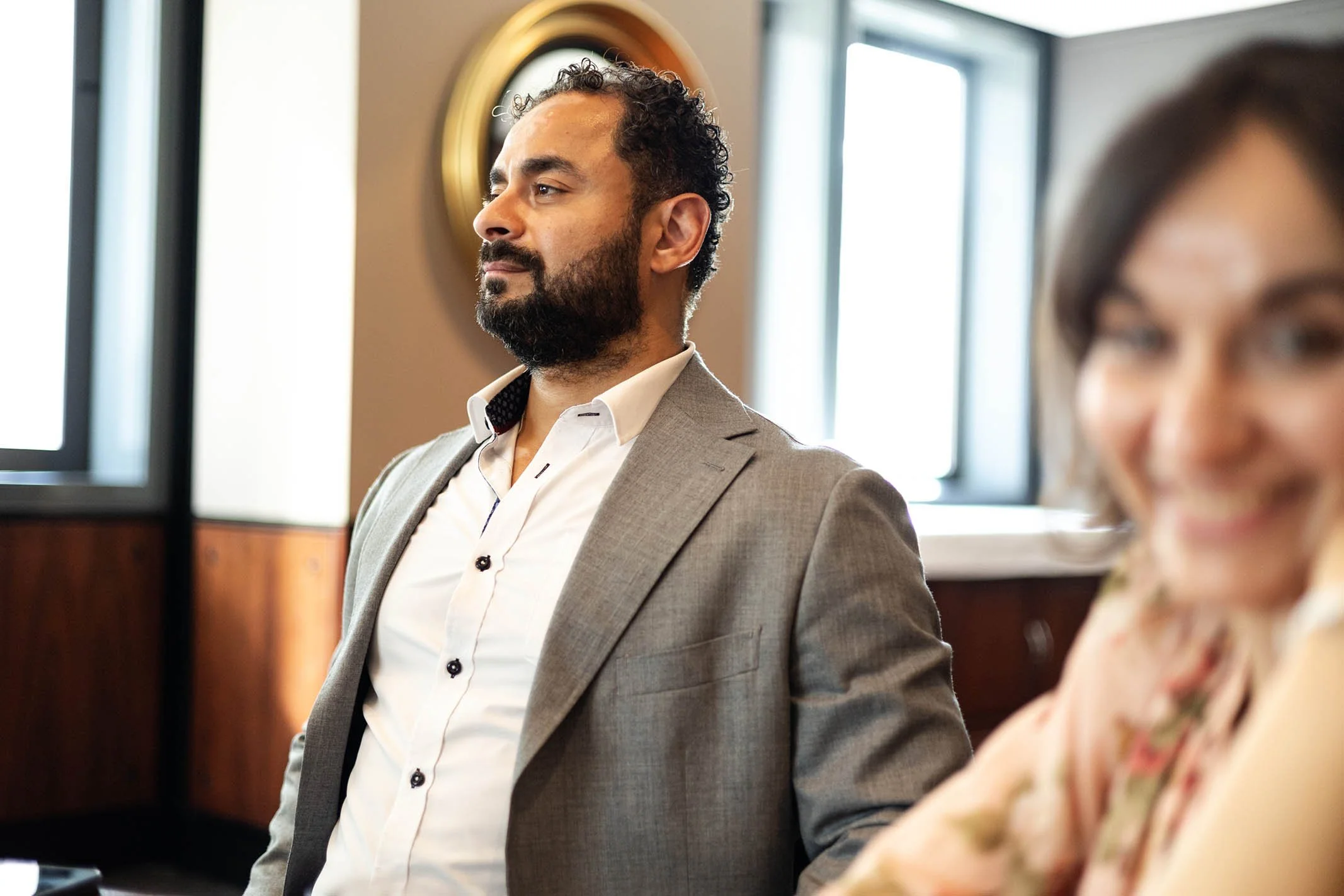 A man in a gray suit and white shirt, with a beard and curly hair, is sitting with a contemplative expression in a room with large windows. A woman with dark hair and glasses, wearing a colorful blouse, is smiling in the foreground, slightly out of f