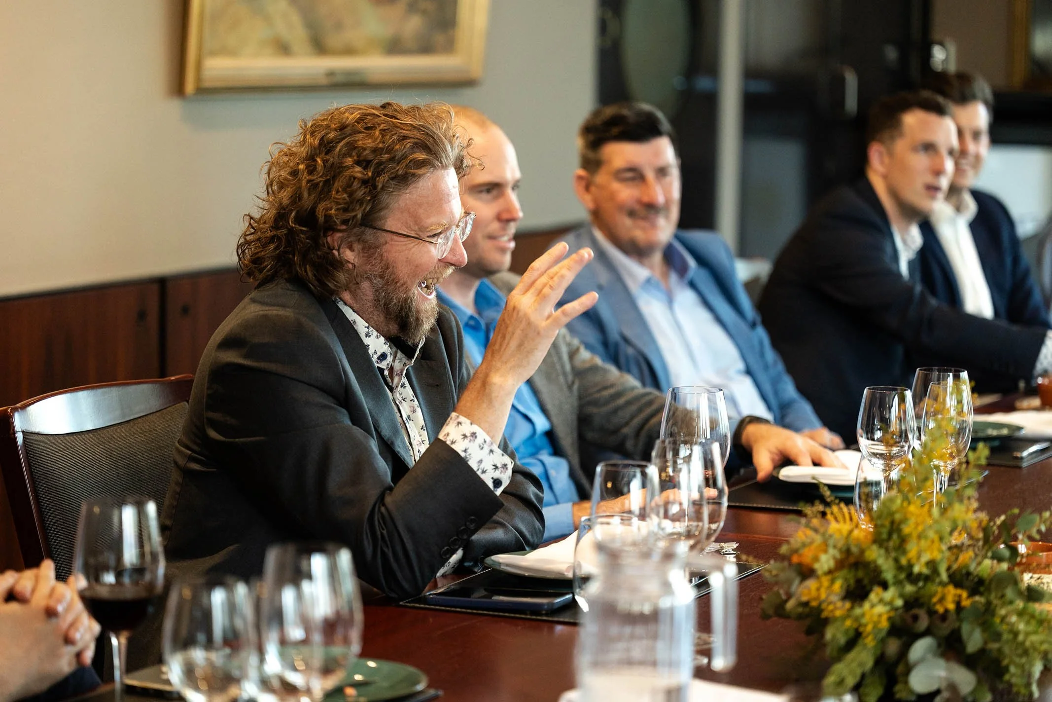 A group of five men in formal attire sitting at a dining table, laughing and engaging in conversation, with wine glasses and a flower centerpiece on the table.