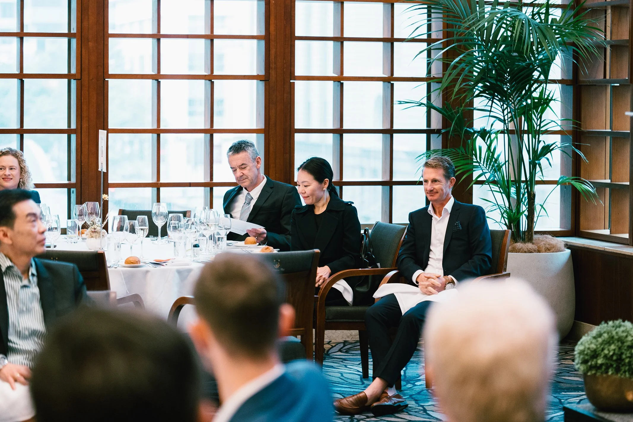 People attending a formal event in a well-lit room with large windows and a wooden grid pattern. Some are seated at a table with glassware and bread, while others are sitting in chairs. A tall potted plant is visible in the background.