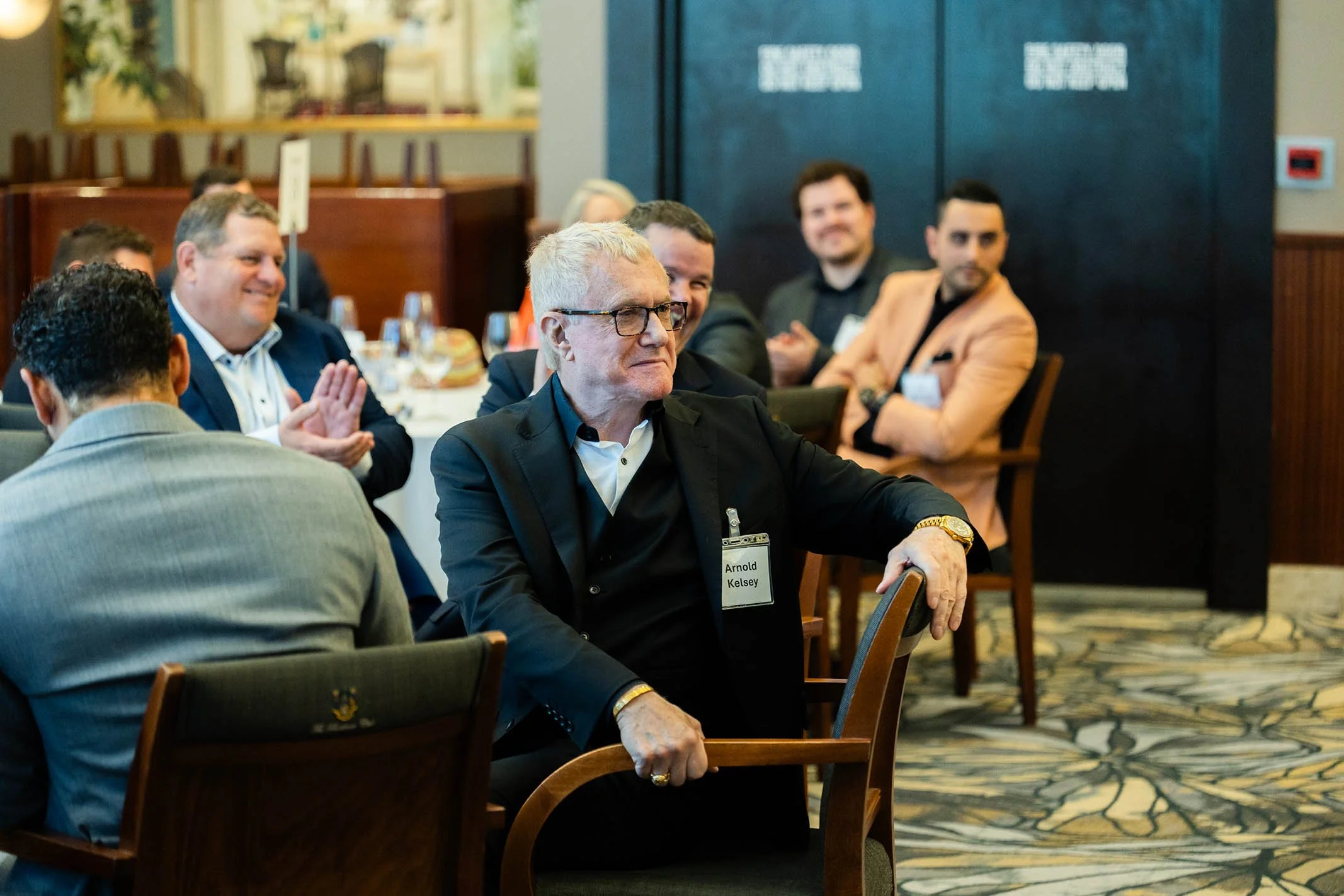 Group of men attending a conference in a restaurant or hotel banquet hall, with one man in the foreground wearing glasses and a black suit, and others clapping or smiling in the background.
