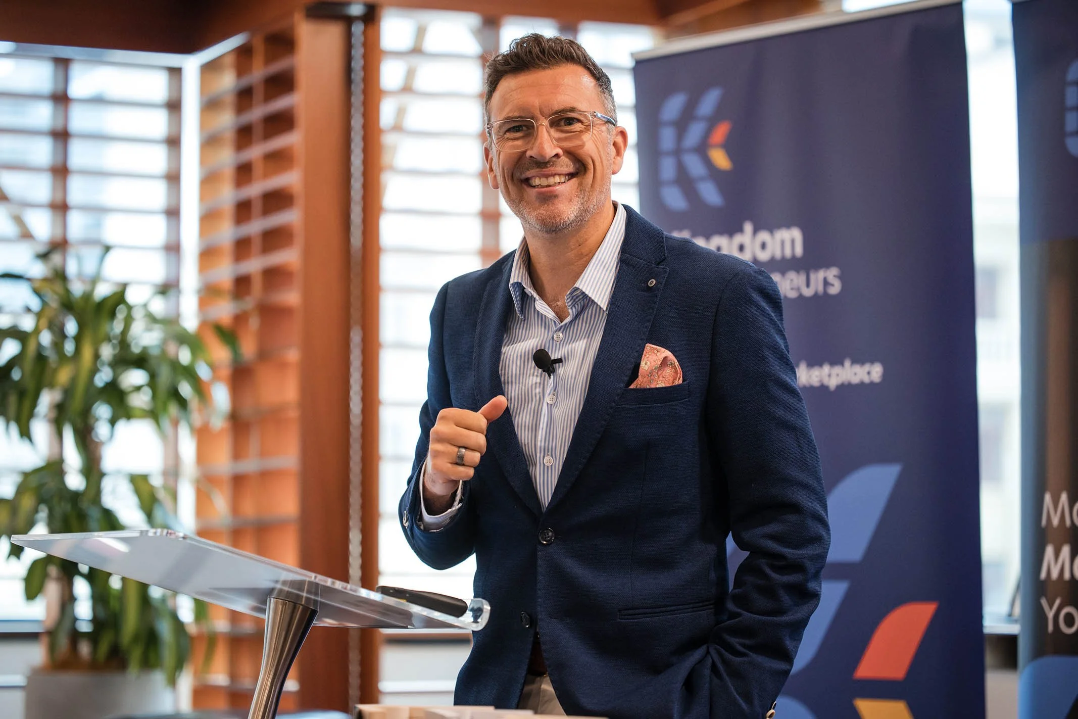 A man in a blue blazer and striped shirt smiling while giving a presentation at a conference.