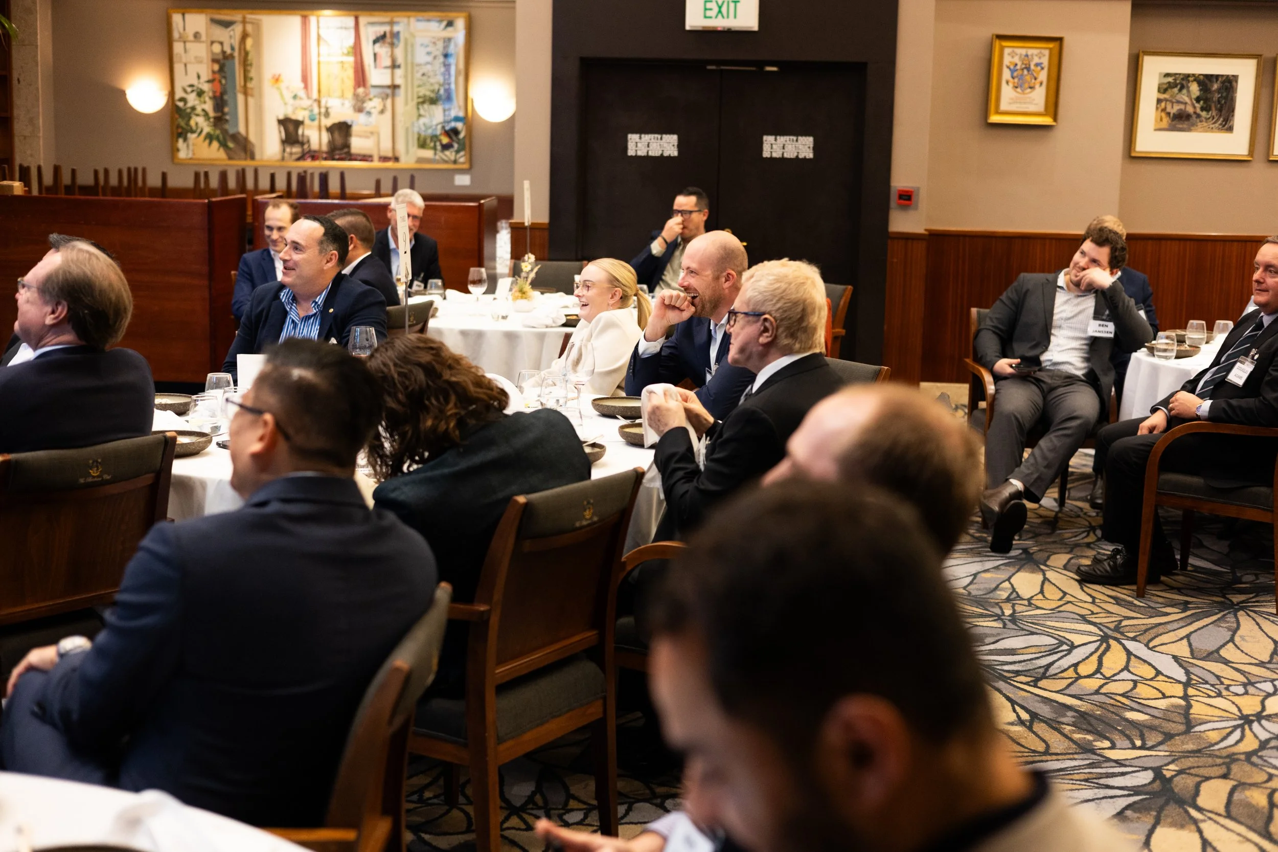 People sitting at round tables in a formal setting, attending a seminar or conference, with some smiling or engaged, in a room decorated with artwork and warm lighting.