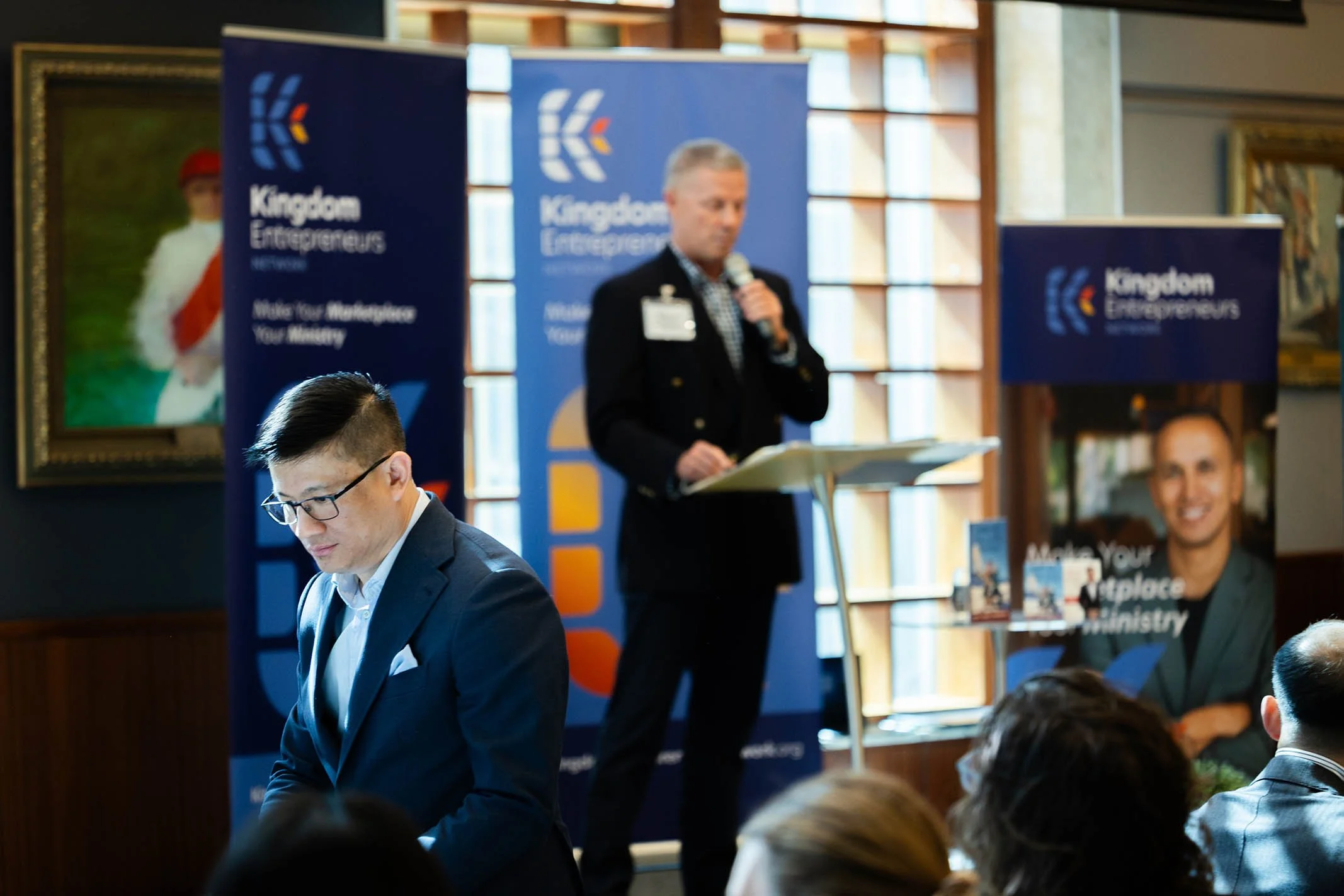 A professional event with a speaker at a podium, holding a microphone, in front of banners that say 'Kingdom Entrepreneurs,' with an audience present. The room has wooden accents and artwork on the walls.