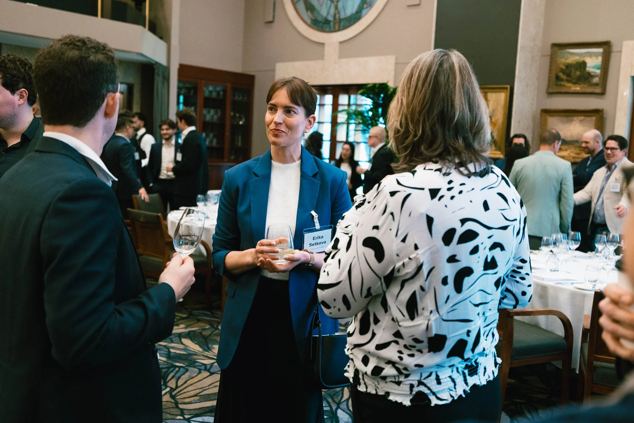 Group of people at a professional networking event, engaging in conversations with glasses of wine or water in hand, in a room with framed artwork and a large window.