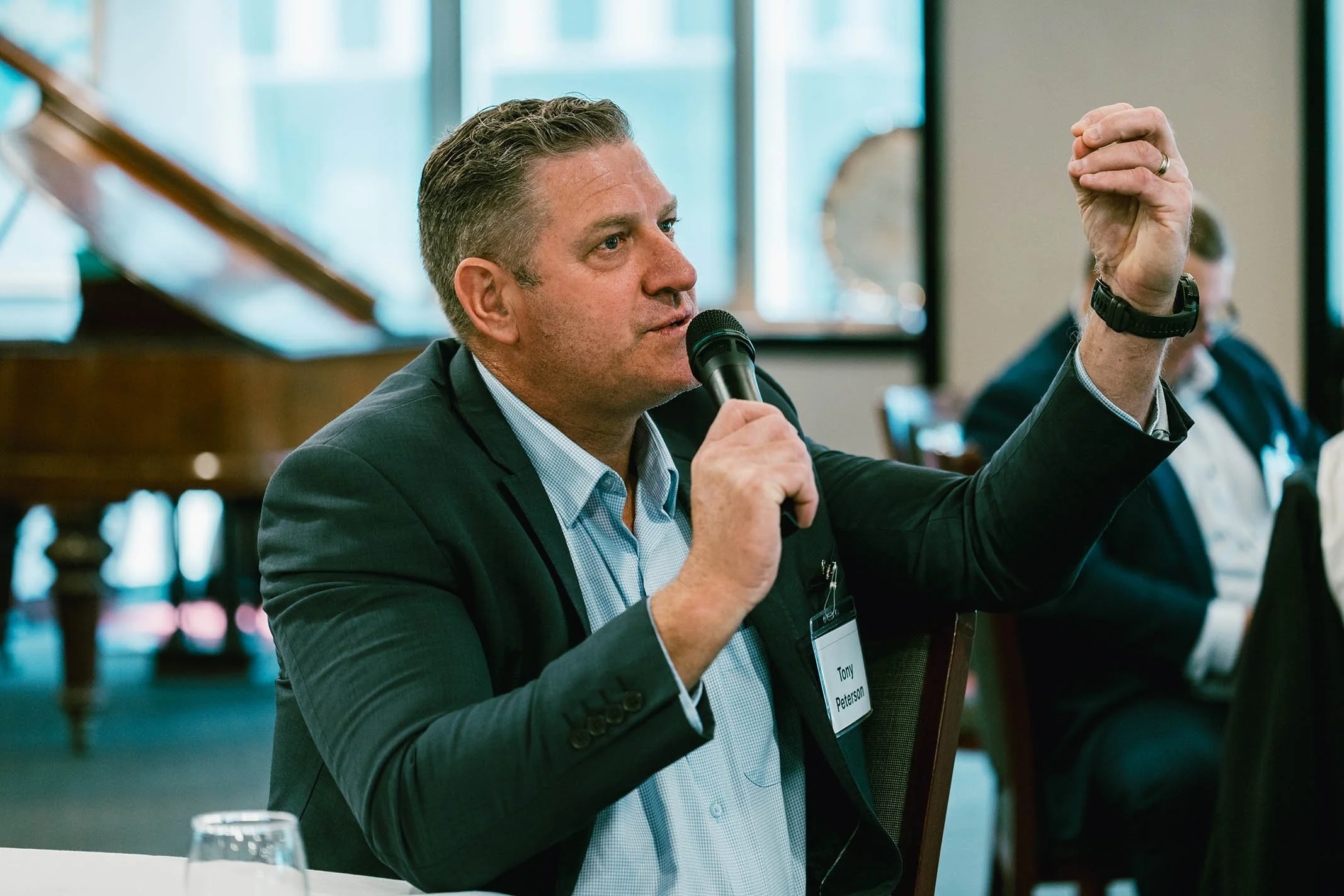 Man in a suit speaking into a microphone at a conference, gesturing with his hand.