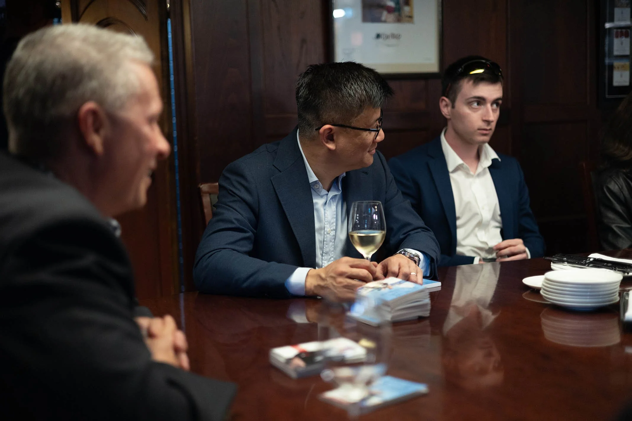 Group of people in business attire sitting around a wooden table in a dimly lit room; one man holds a glass of white wine and looks to his right.