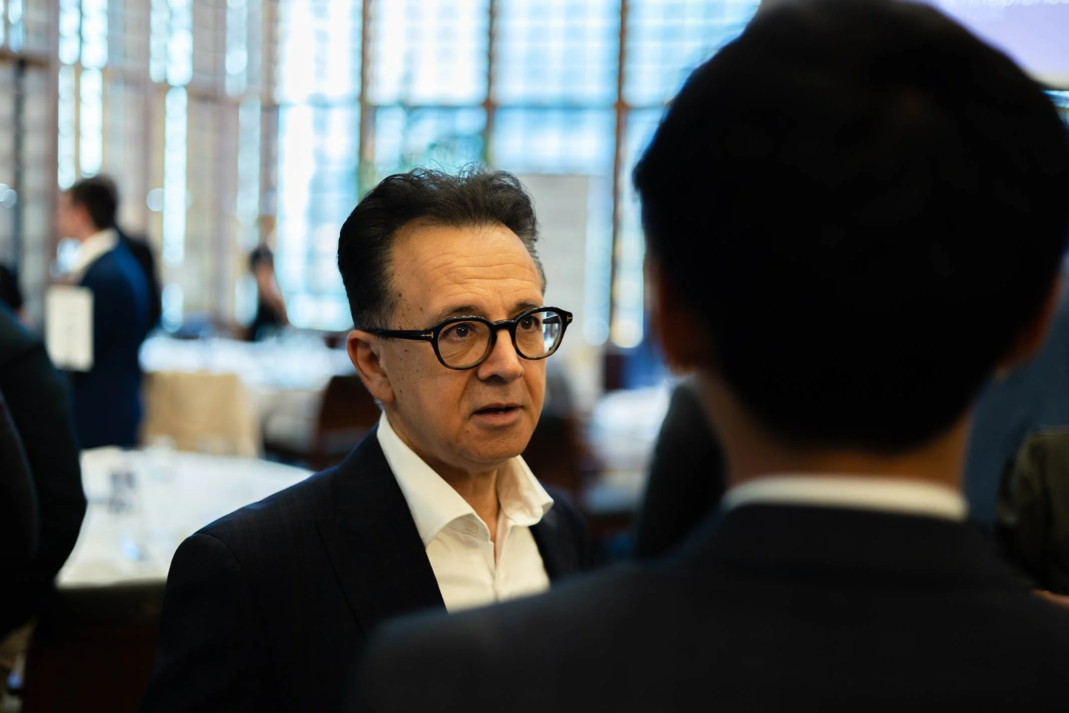 A man wearing glasses and a white shirt is in conversation with another person, with a blurred background of a restaurant or conference setting.
