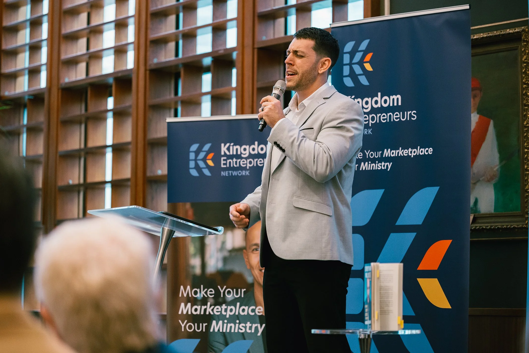 A man in a light gray blazer speaking into a microphone at a speaking event with a blue Kingdom Entrepreneurs banner behind him.